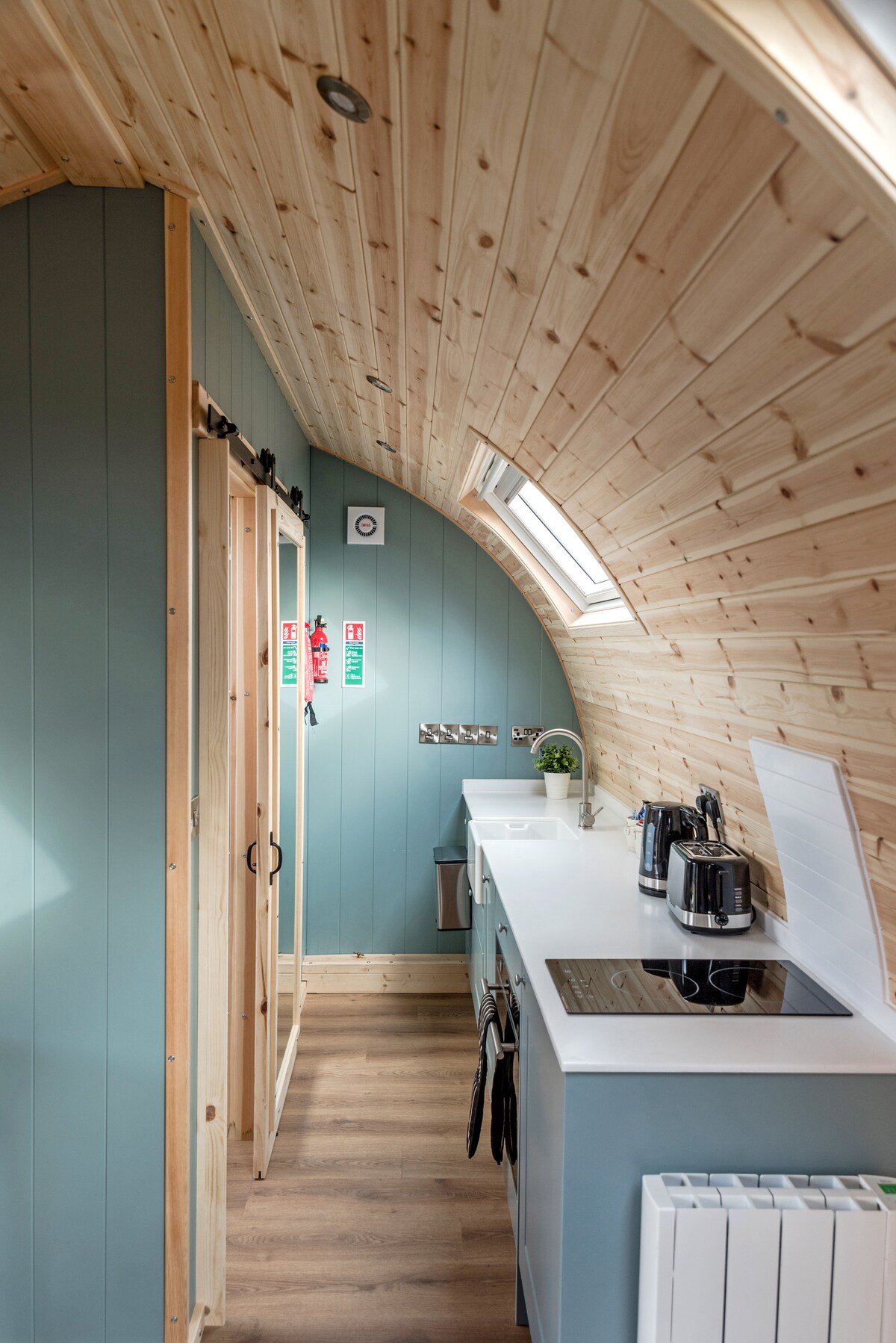 An interior view of a glamping pod features an arched wooden ceiling and blue-painted walls. A well-equipped kitchen area is visible, showcasing a stove, kettle, and storage elements. A small window allows natural light to illuminate the space, enhancing the inviting atmosphere.