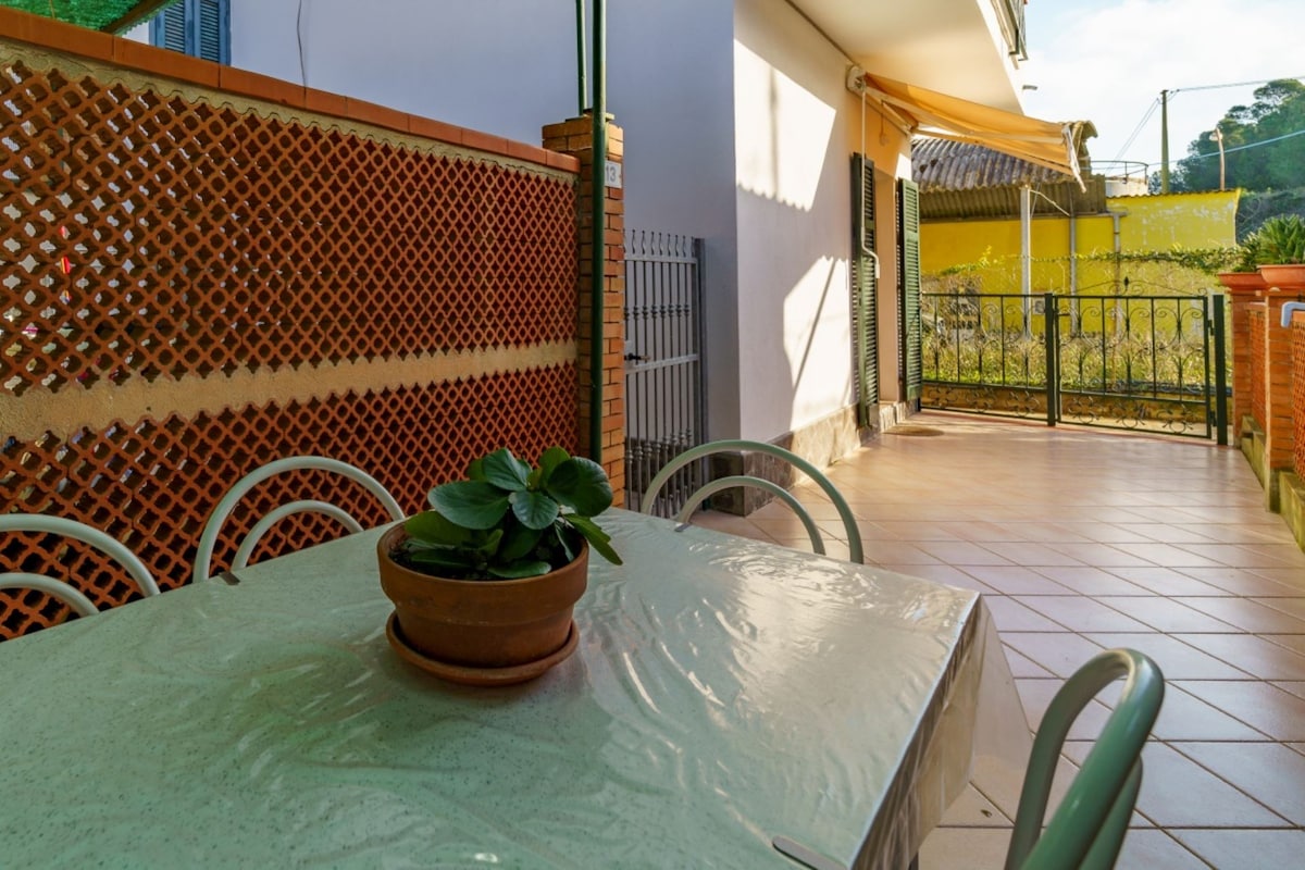 An outdoor area is presented with a covered dining table set, featuring a plant in a terracotta pot as the centerpiece. The tiled floor reflects natural light, while a fence provides privacy. A view of the surrounding greenery is visible in the background.