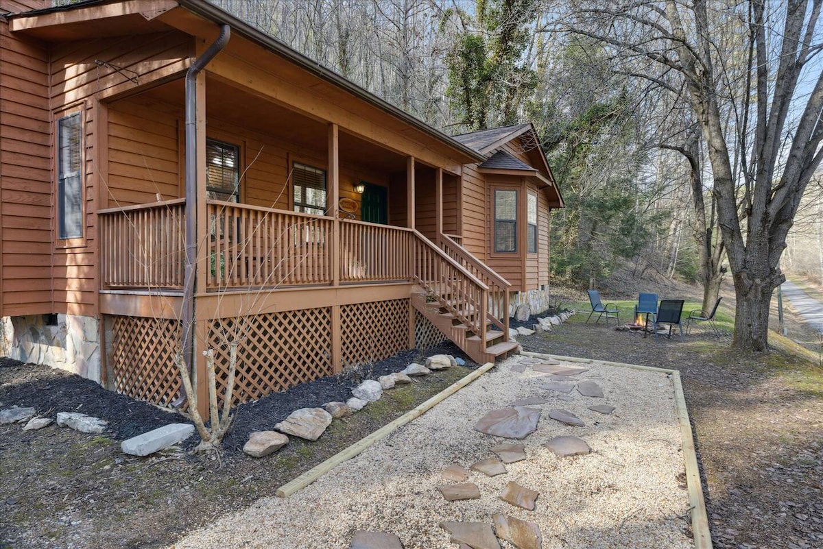 The exterior of a wooden cabin is showcased, featuring a wraparound porch with a railing and steps leading to the entrance. A stone pathway is visible, bordered by gravel and accentuated with natural landscaping. Chairs are arranged in a small seating area nearby.