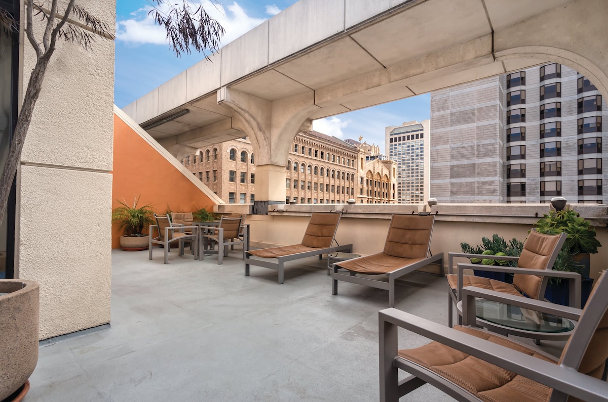 A spacious rooftop sun deck is featured, furnished with several lounge chairs arranged for relaxation. Potted plants add greenery to the area, while the background highlights the urban architecture of San Francisco's buildings under a clear blue sky.
