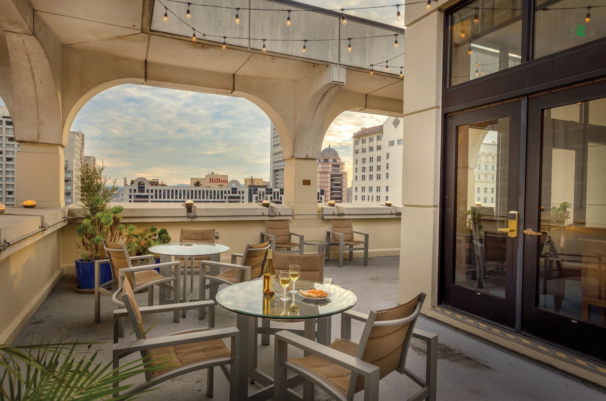 An outdoor sun deck features several tables and chairs, offering a relaxed seating area with a view of the city. String lights hang above, providing a warm ambiance. Potted plants add greenery to the space, enhancing the rooftop setting.