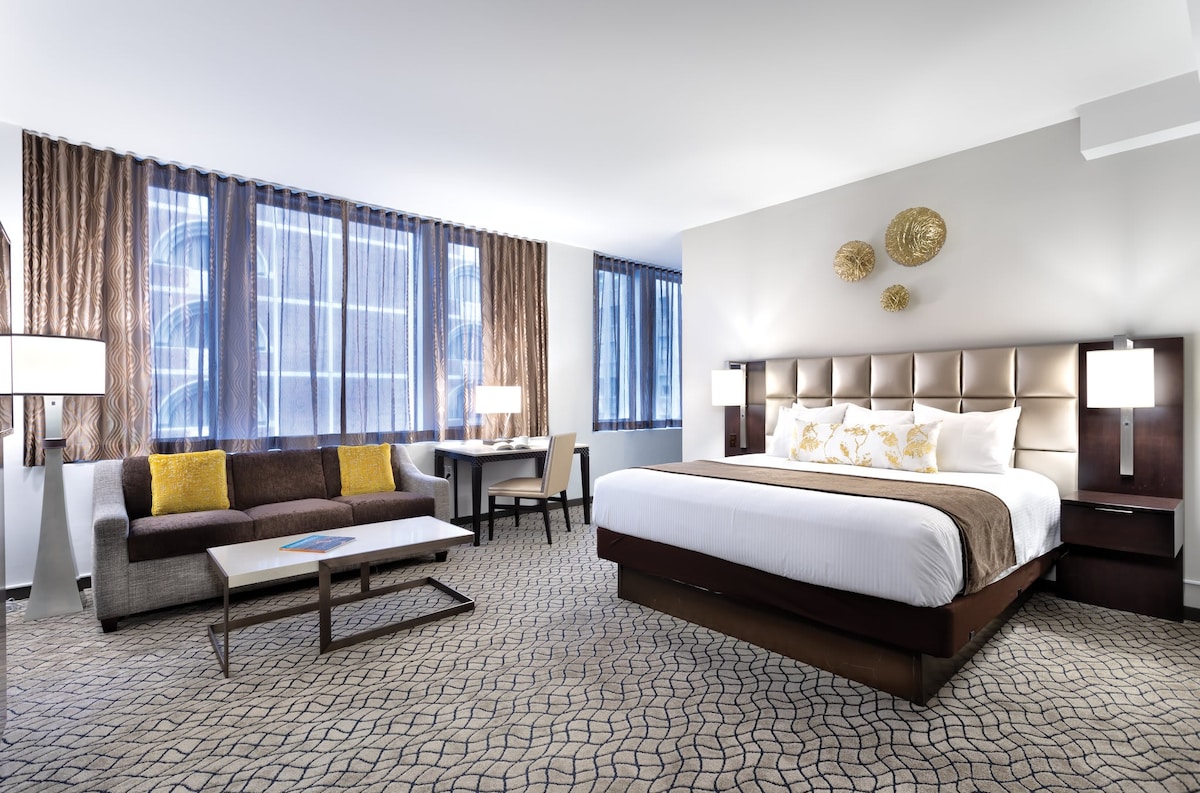 A spacious room features a large bed with decorative pillows and a stylish headboard, complemented by a cozy sofa and a coffee table. Natural light filters through floor-to-ceiling windows, and modern artwork adorns the walls, creating a serene atmosphere for relaxation.
