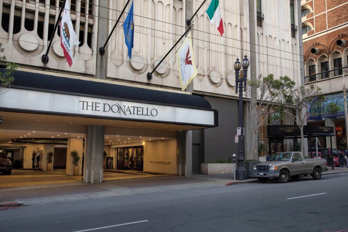 The exterior of The Donatello is prominently displayed, showcasing an entrance under a covered awning. Flags representing California, the United States, and Italy are visible. A street lined with trees and parked cars is adjacent to the entrance, contributing to the urban atmosphere.