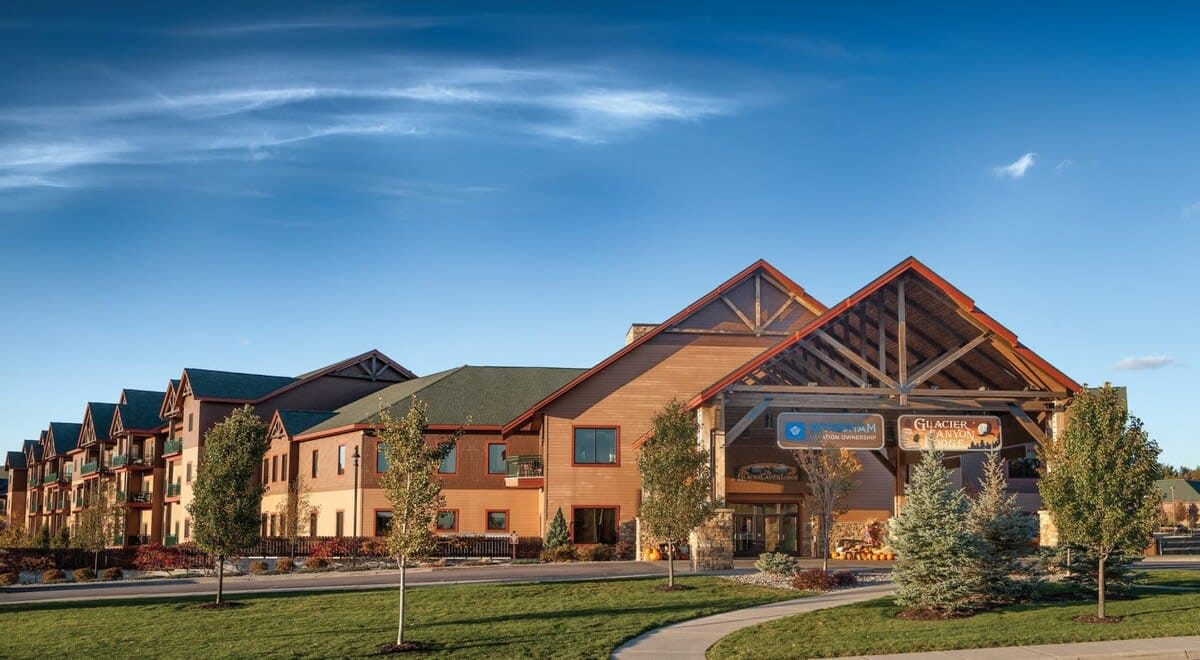 The resort's exterior features a blend of modern and rustic architecture, highlighted by timber accents and stone details. Lush landscaping and well-maintained grass areas surround the entrance, creating an inviting approach. The clear blue sky complements the serene outdoor setting.