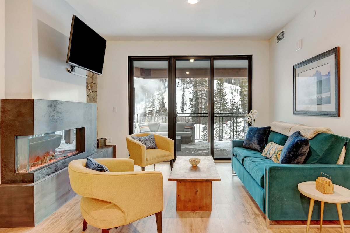 The living area showcases a turquoise sofa paired with two armchairs, offering a cozy spot beside a modern gas fireplace. Large glass doors provide a view of the snowy landscape outside, while a wooden coffee table centers the space.