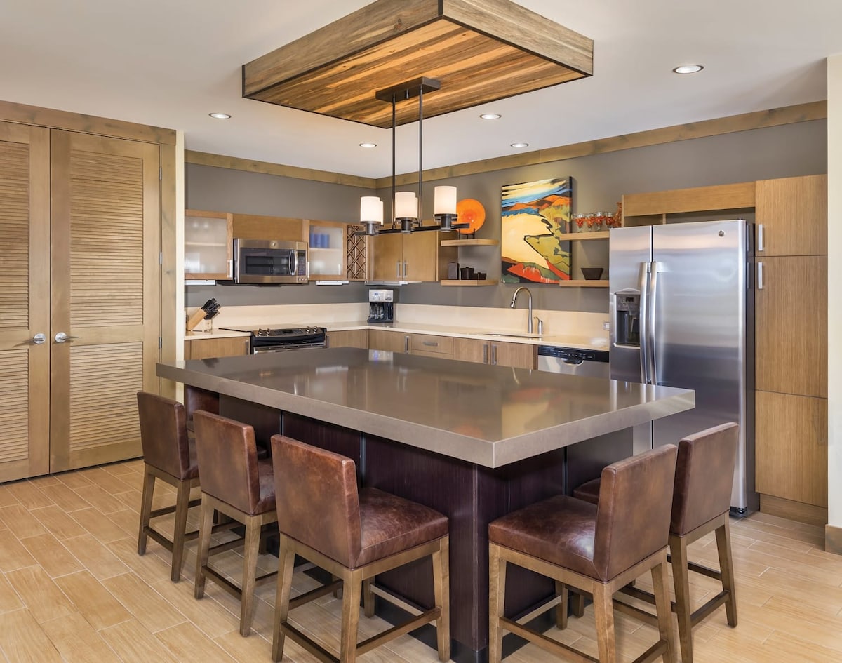 A modern kitchen area features a large rectangular island with a polished countertop, surrounded by six textured brown chairs. Wooden cabinetry complements stainless steel appliances and a colorful piece of artwork adds a touch of vibrancy to the neutral color scheme.