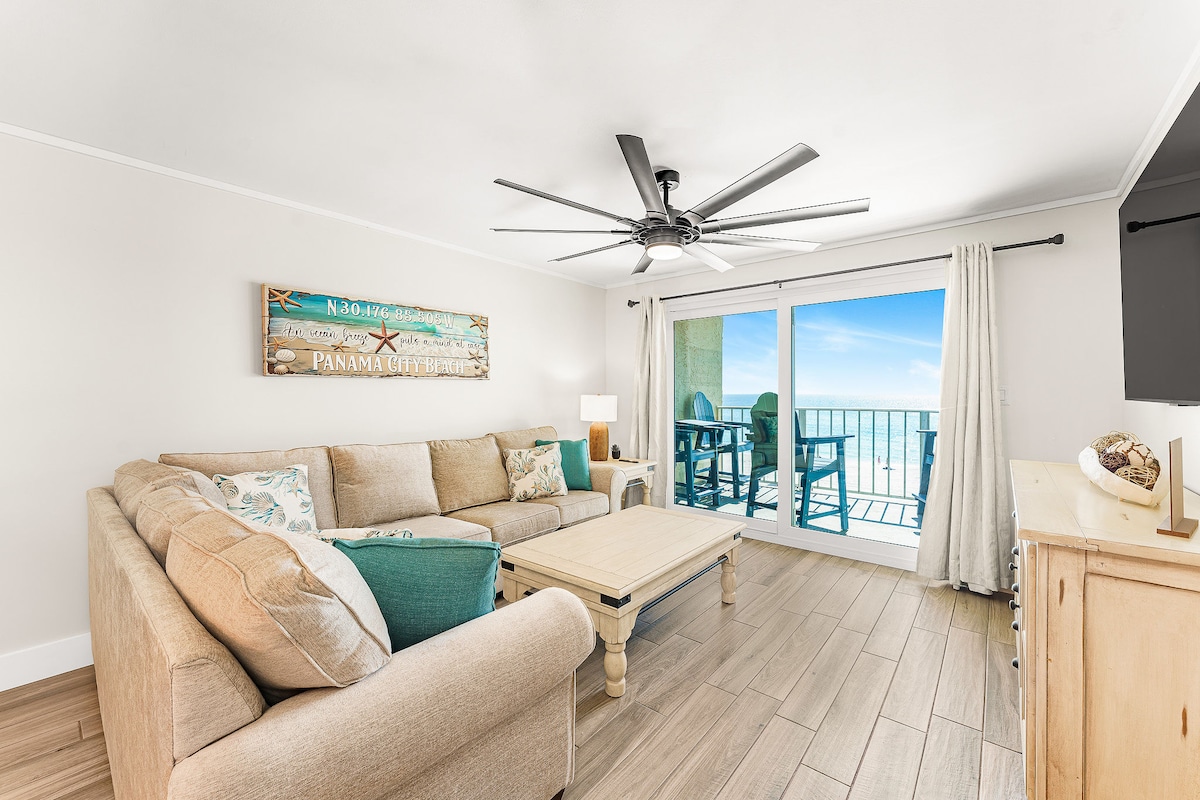 A spacious living area is furnished with a light-colored sectional sofa and a coffee table. Large sliding glass doors provide access to the balcony and showcase panoramic ocean views. A ceiling fan and soft lighting add to the bright, open atmosphere of the room.