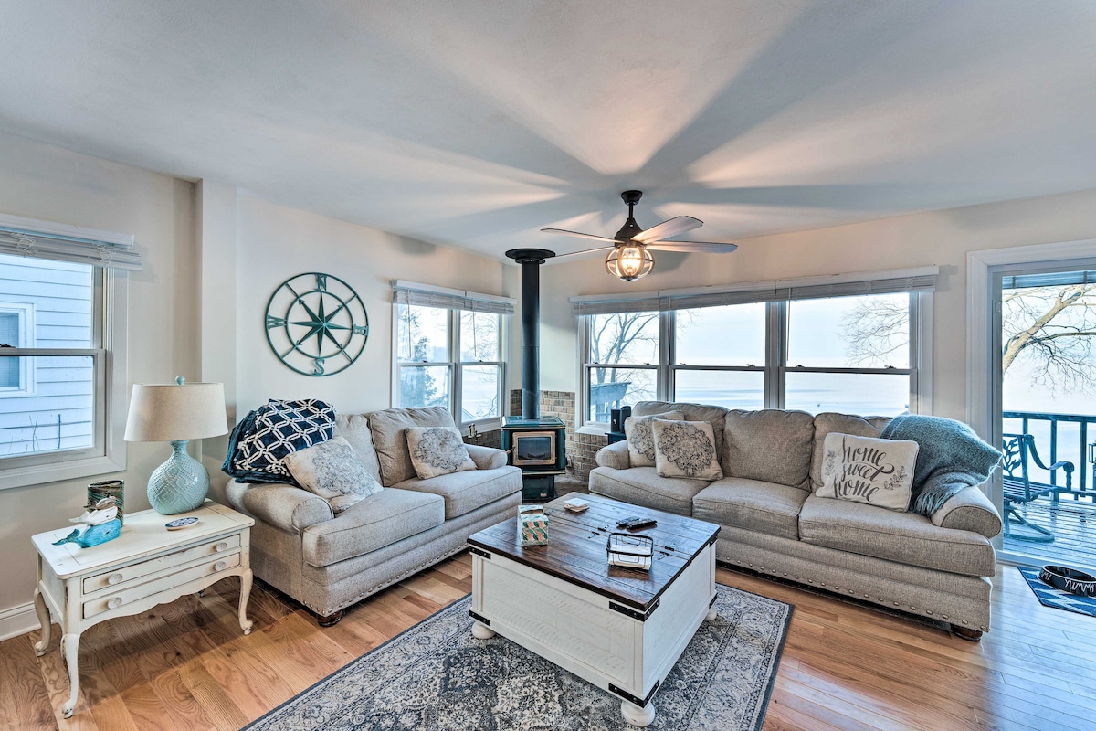 A bright living area features two comfortable sofas facing each other, offering a relaxed seating arrangement. Large windows provide views of Lake Koshkonong, while a decorative area rug adds warmth to the hardwood floor. A ceiling fan and a stylish light fixture enhance the space.