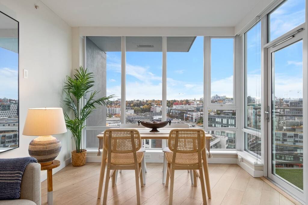 A bright dining area features a wooden table surrounded by two woven chairs, offering a view through large glass windows. A decorative plant is positioned beside the table, and a softly glowing lamp adds warmth to the space. The scenery outside showcases an urban landscape.