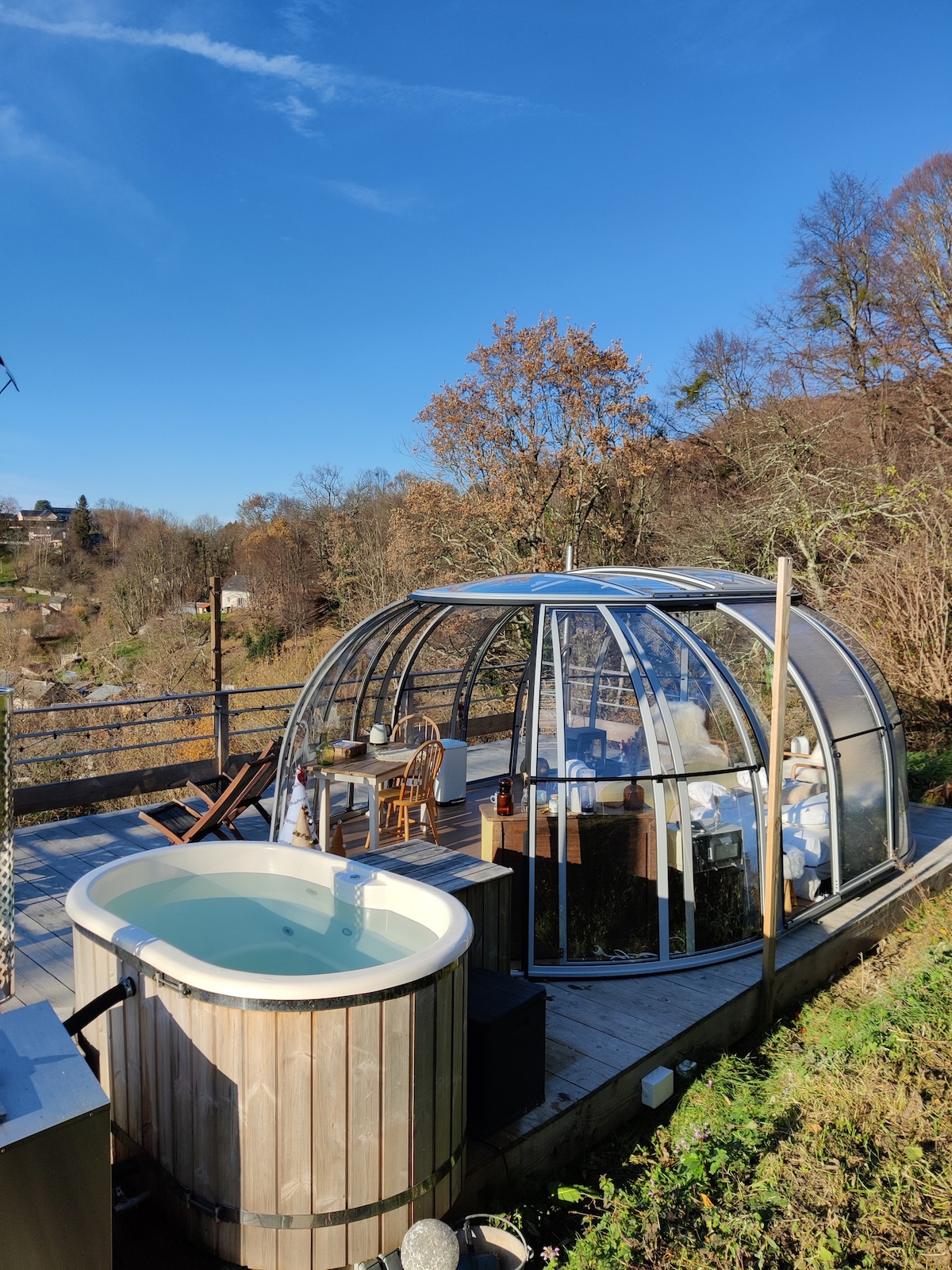 A private terrace features a wooden hot tub surrounded by greenery. The glass dome structure offers a spacious interior illuminated by natural light, showcasing a dining area and seating arrangements. Clear blue skies and seasonal foliage provide a serene backdrop.