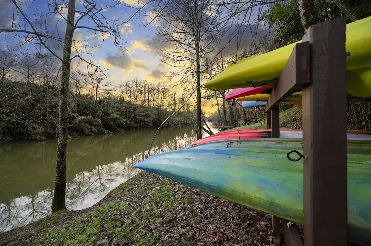Colorful kayaks are stored on a wooden rack beside a calm river, reflecting the vibrant hues against the tranquil water. Leafless trees surround the area, with an evening sky showcasing soft clouds and warm tones as the sun sets in the distance.
