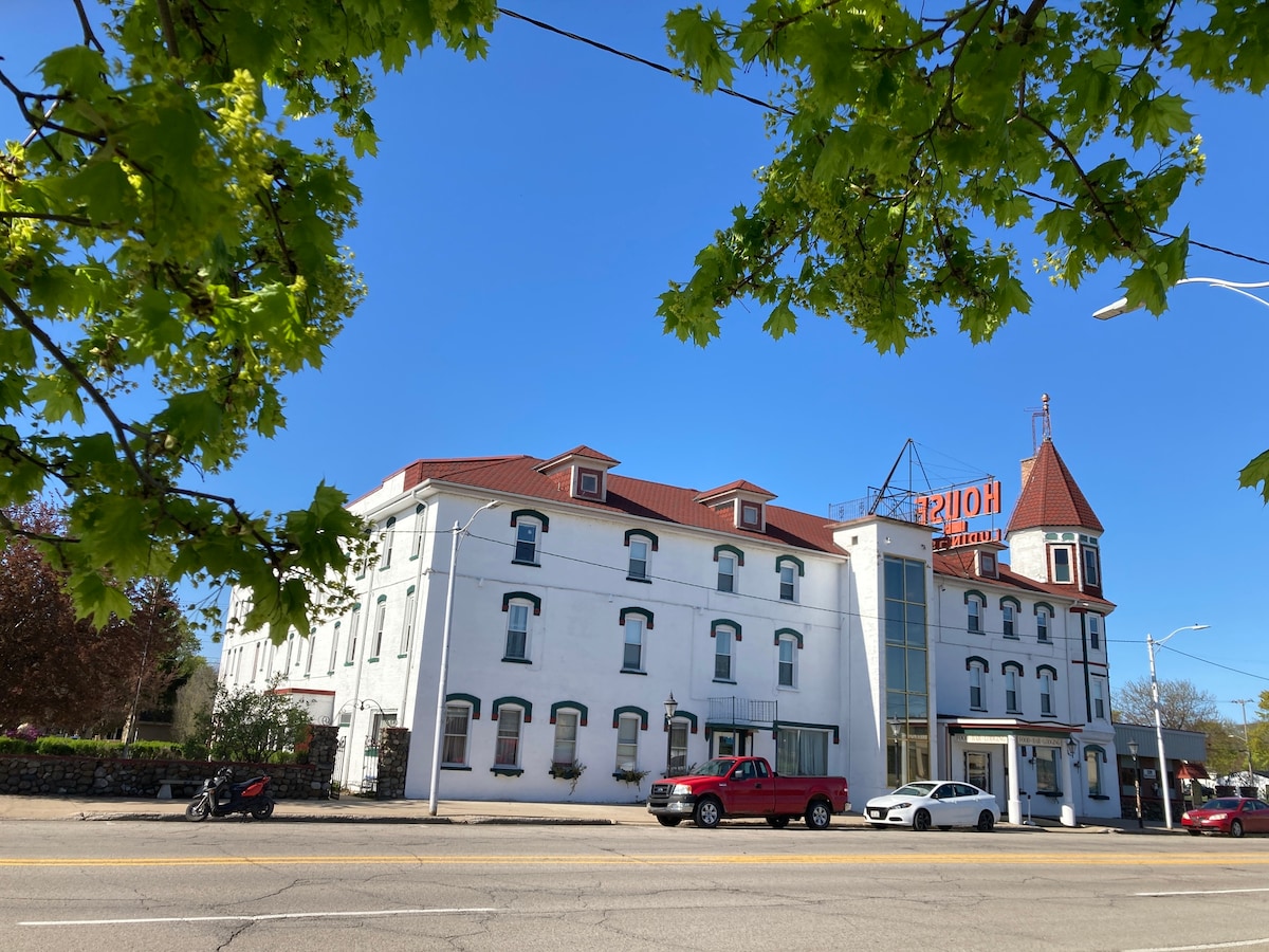 The exterior of the Historic House of Ludington is displayed, featuring a three-story white building with a distinct tower. A clear blue sky serves as the backdrop, while nearby trees provide a touch of green. Vehicles are parked in front, enhancing the bustling atmosphere.