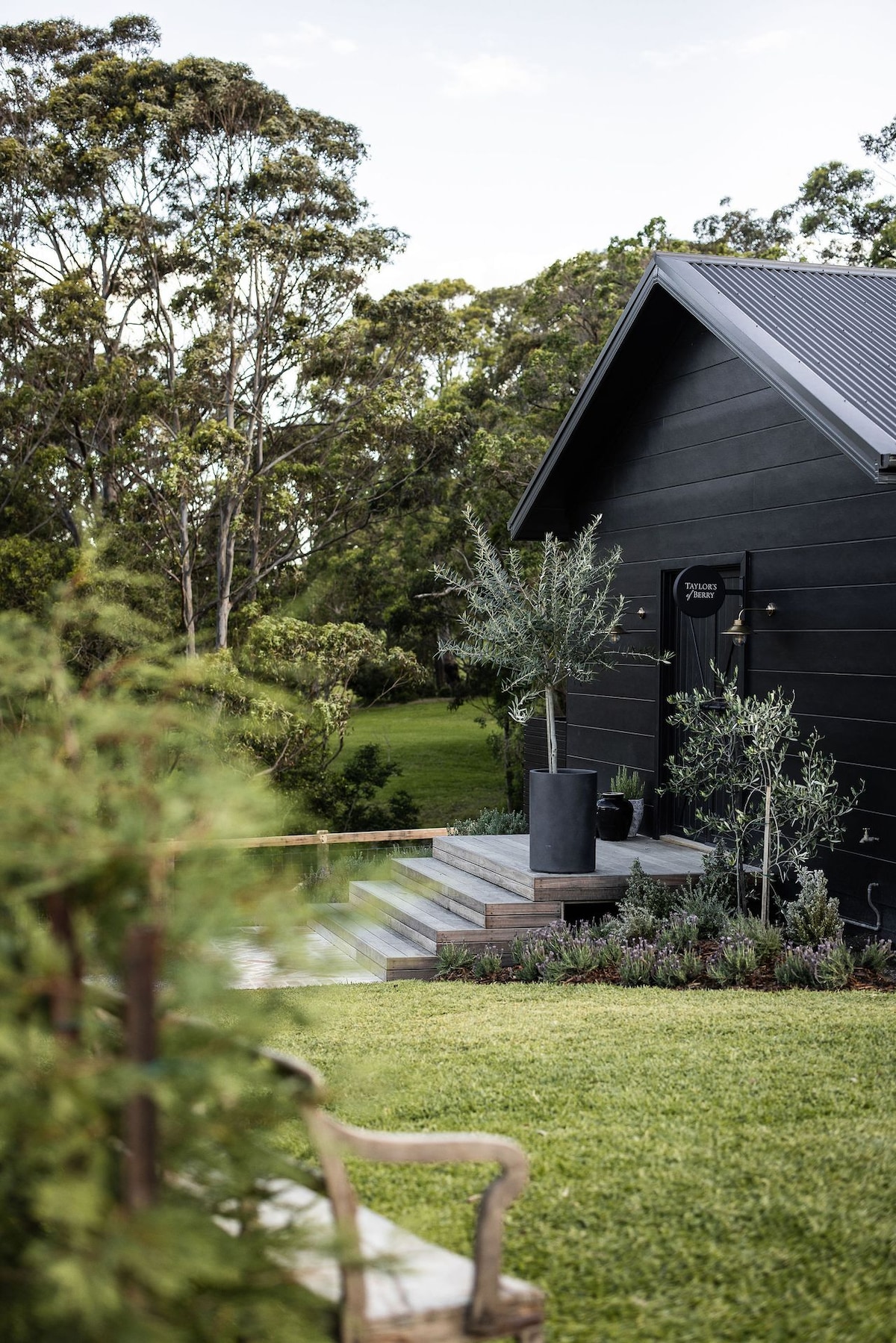 The exterior of the guest house is highlighted by a dark timber-clad facade. Steps lead up to a stylish front door, surrounded by lush greenery and ornamental plants. A mix of flowering shrubs and manicured grass create a welcoming atmosphere.