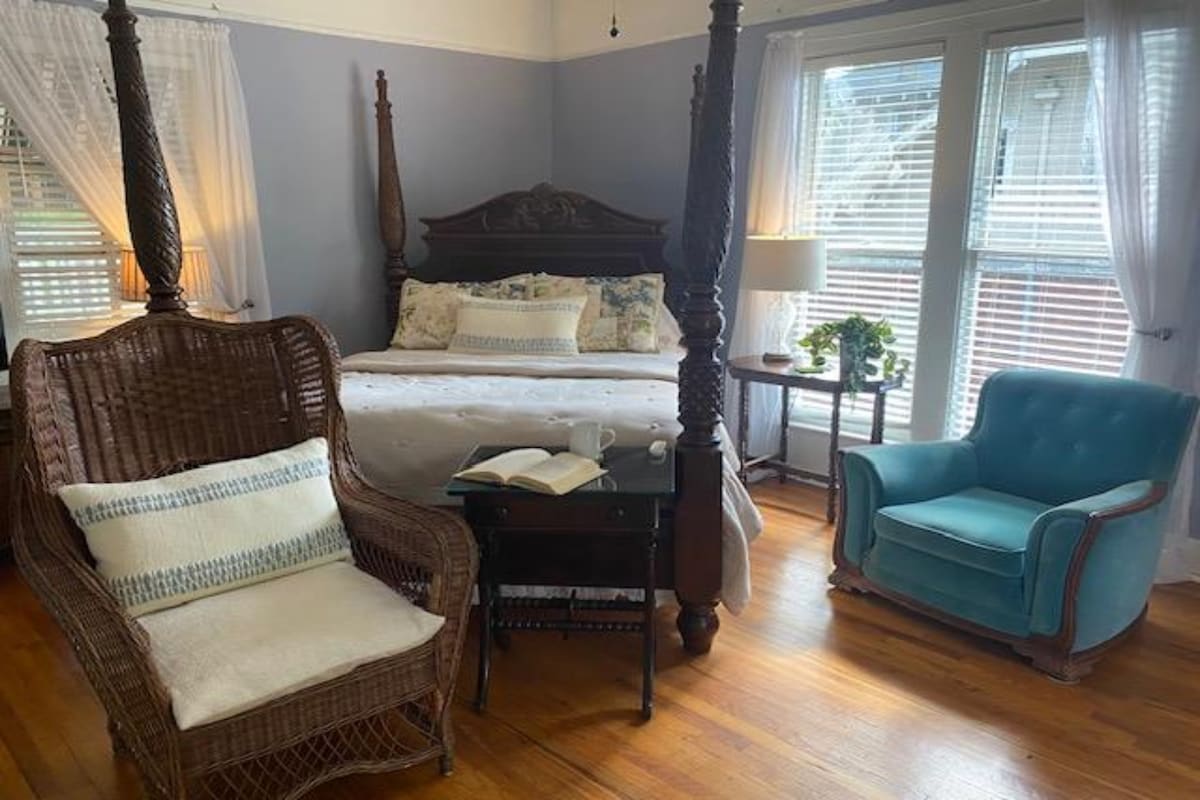 A spacious bedroom is highlighted by a large four-poster bed with decorative bedding. A wicker chair and a blue armchair provide comfortable seating options. Natural light filters through the window, illuminating the warm wooden floor and small side table with books and cups.