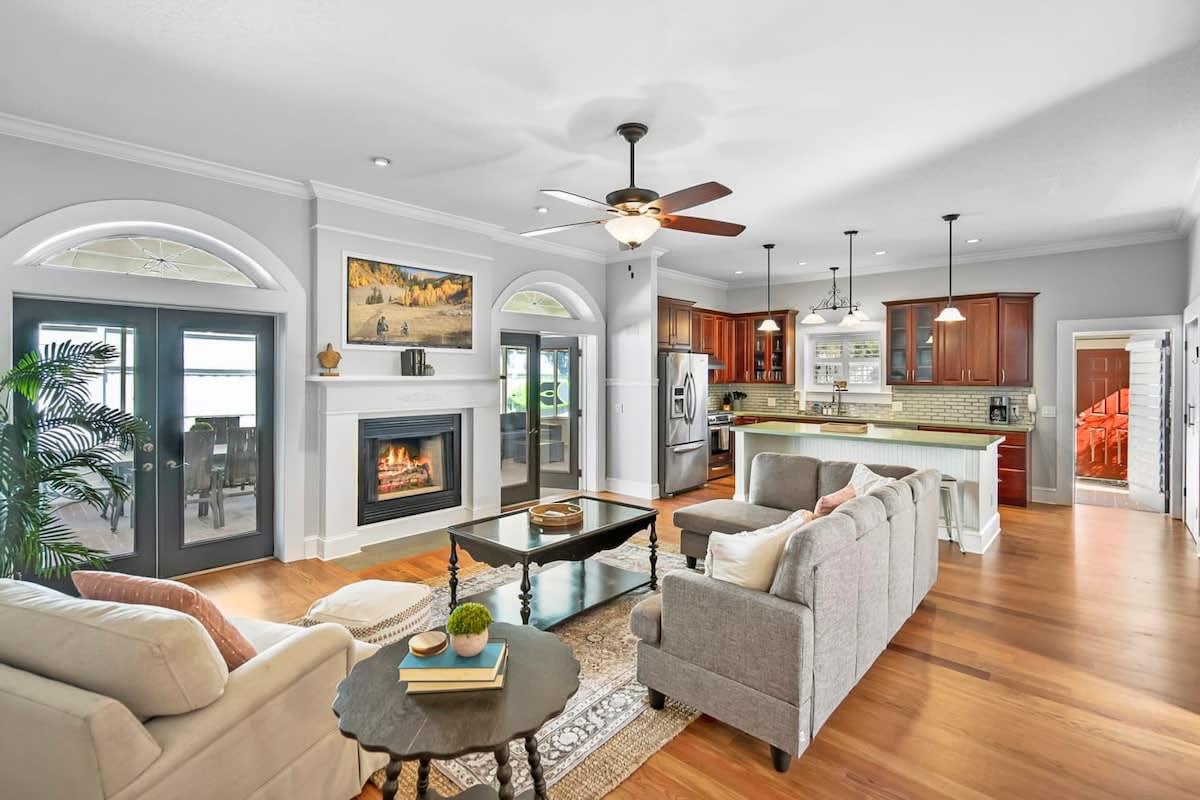 A spacious living area features wood flooring and a cozy fireplace. Comfortable seating includes a beige sofa and gray armchairs arranged around a dark coffee table. Large windows provide natural light, and an open kitchen showcases wood cabinetry and modern appliances in the background.