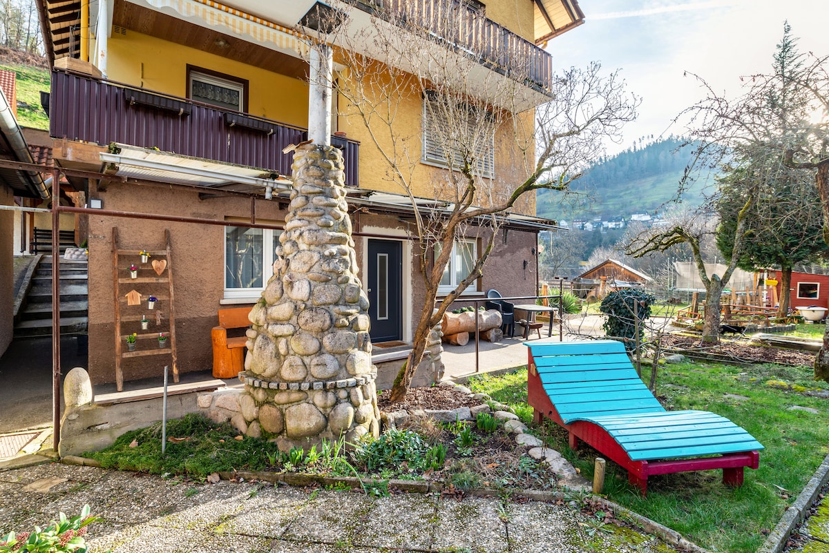 The outdoor area features a garden space with a unique stone feature and colorful lounge chairs. A natural stone barbecue is visible alongside a wooden seating area. Nearby, a tree and greenery enhance the inviting outdoor setting with views of the surrounding hills.