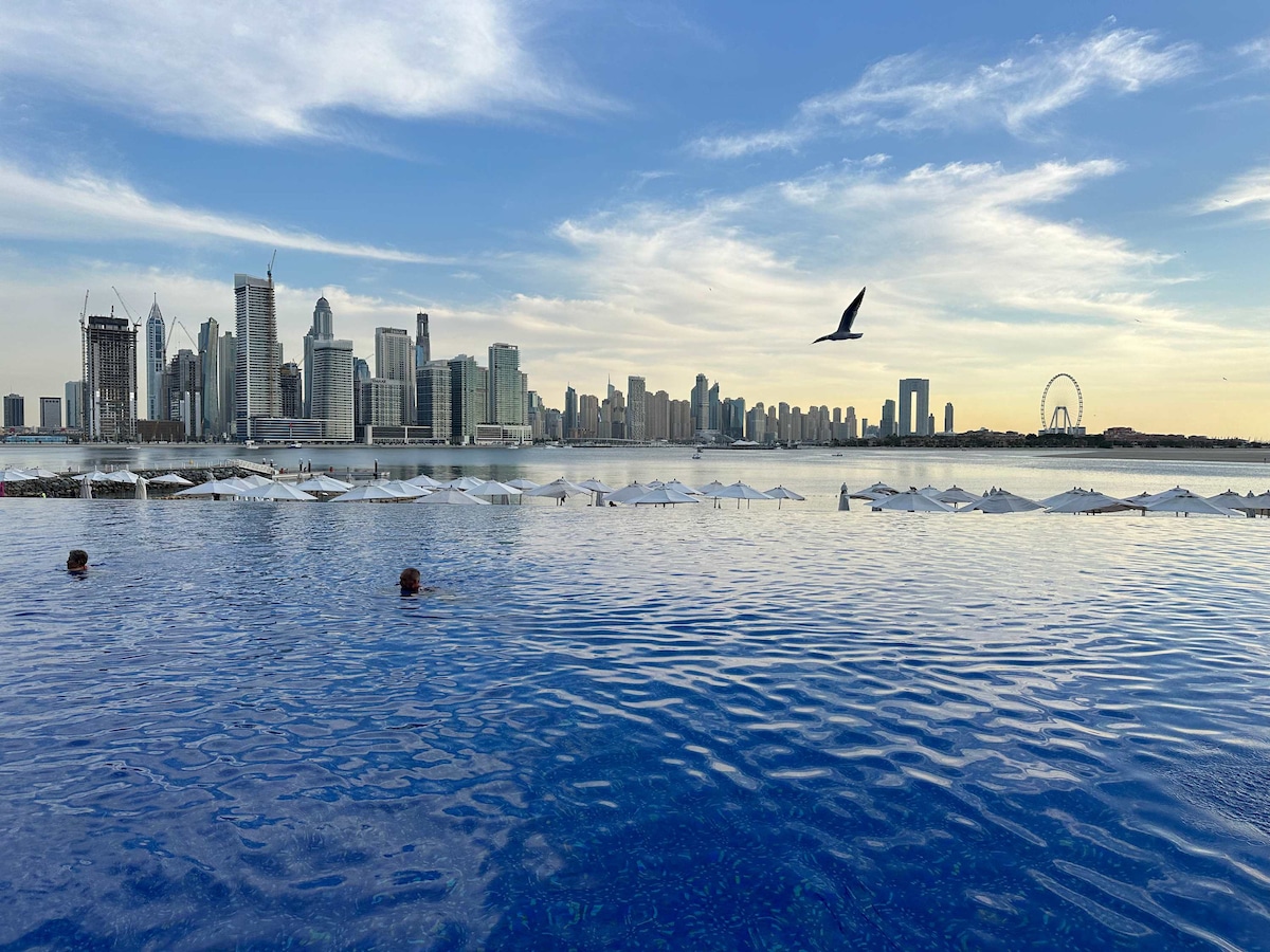 An infinity pool reflects the skyline of Dubai, featuring modern skyscrapers and architectural landmarks. Lounge chairs are visible along the poolside, and umbrellas provide shade. A clear blue sky is present, with a silhouette of a bird flying overhead.