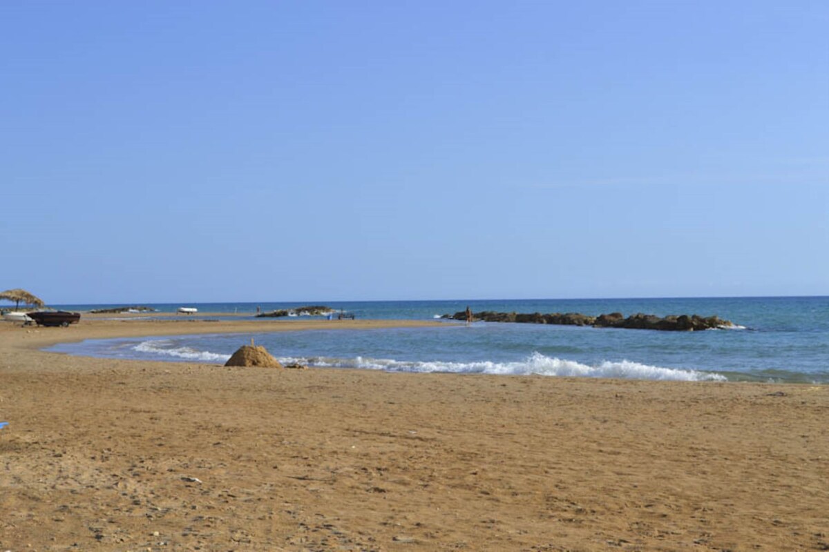 A sandy beach stretches toward the horizon, meeting the gentle waves of the Mediterranean Sea. The calm water reflects the clear blue sky, while natural elements like rocks and a distant boat add to the coastal charm. Sunlight bathes the landscape in a warm glow.