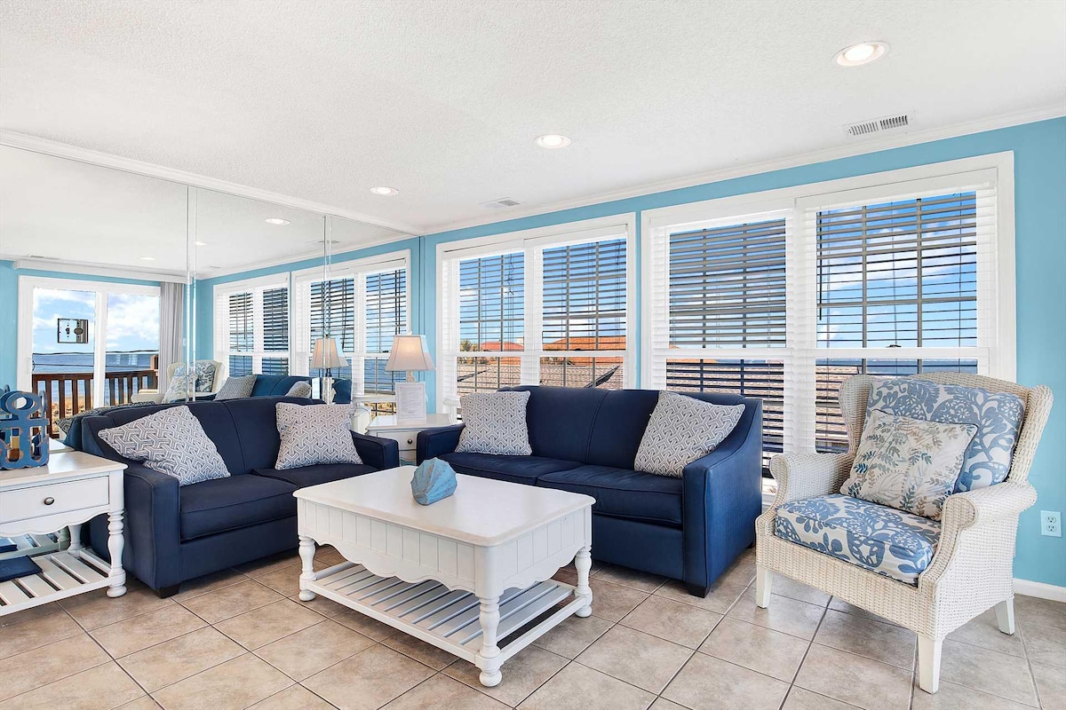 The living room features ocean views through large windows, with natural light creating a bright atmosphere. Two navy blue sofas and a white coffee table are arranged on tiled flooring. A light-colored armchair and a console table complement the space, which is decorated with subtle coastal accents.