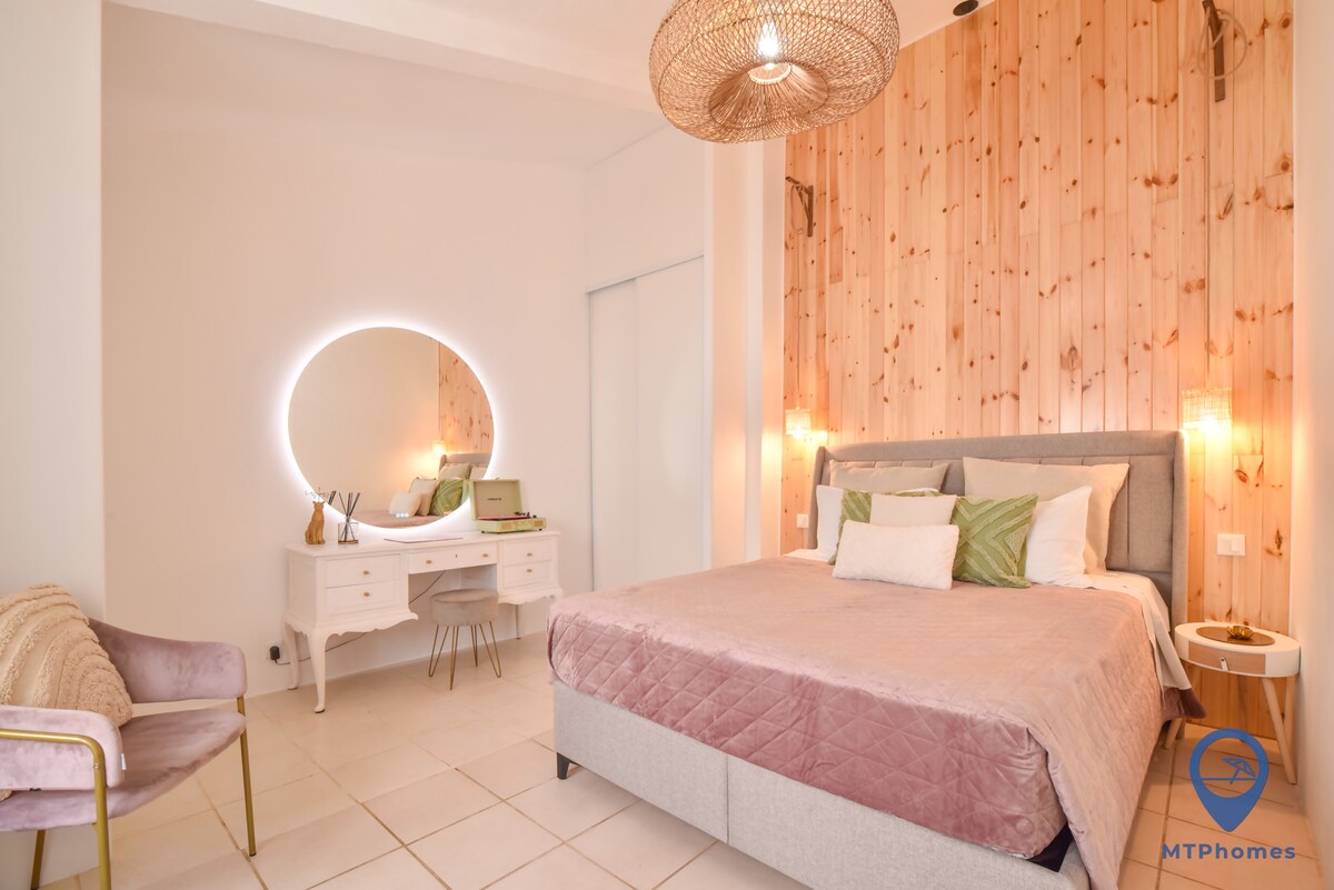 A well-lit bedroom features a spacious bed with plush bedding and decorative pillows. A mirrored vanity is positioned near the bed, complemented by stylish lighting. Natural wood paneling adds warmth to the room, while soft colors create a calming atmosphere.