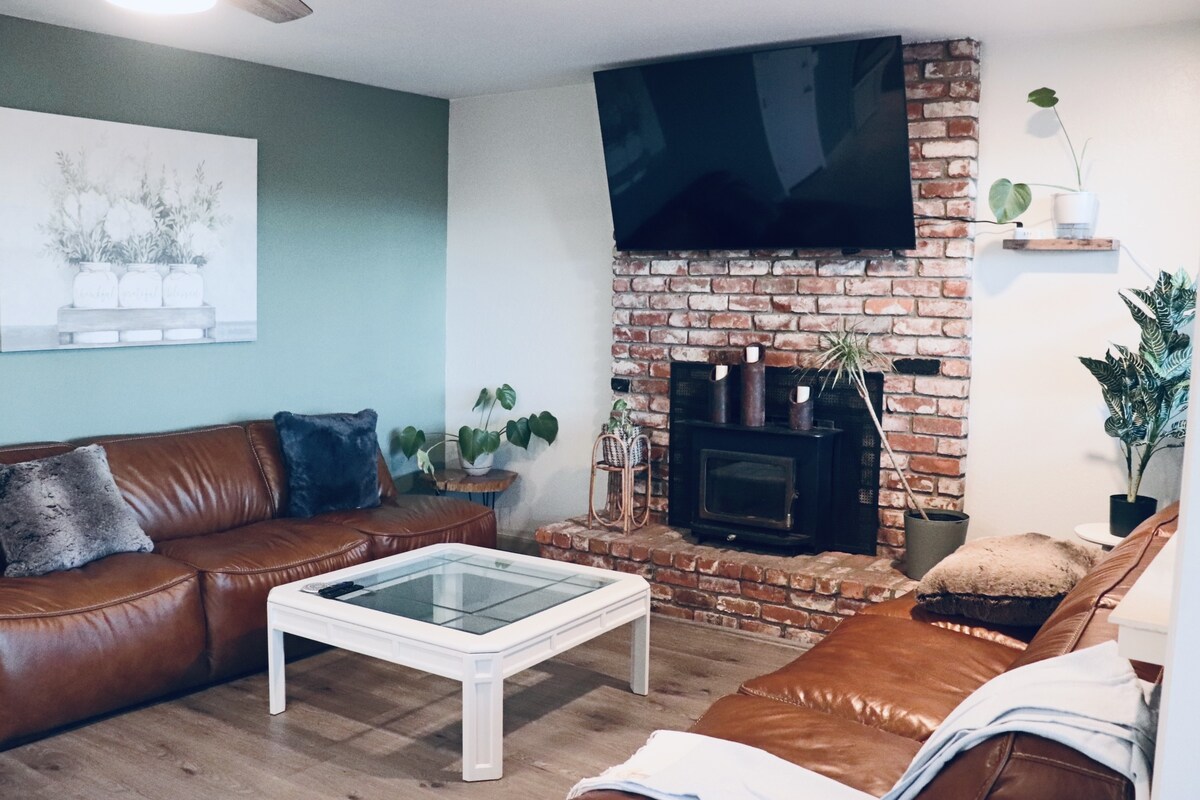 A cozy living room features a mix of leather sofas and a glass coffee table, inviting relaxation. A brick fireplace serves as a focal point, surrounded by plants and decorative elements. A mounted television is positioned above the fireplace, enhancing the comfortable ambiance.