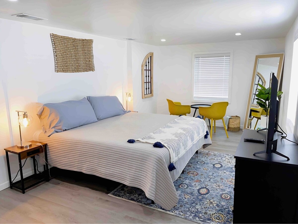 A bright sleeping area features a spacious bed with light blue bedding and two bedside lamps. A patterned rug enhances the floor, while a mirror and a small dining table with yellow chairs are positioned nearby. Natural light filters through the window, illuminating the space.