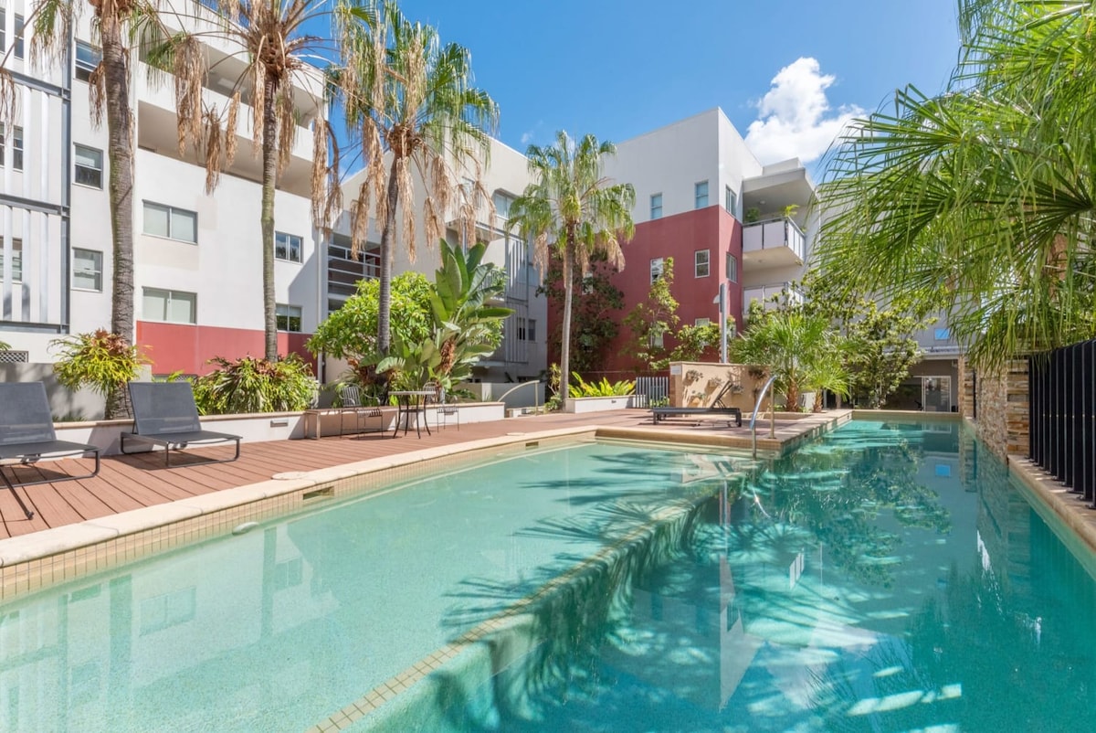 The image displays a serene pool area surrounded by lush greenery and palm trees. Lounge chairs are positioned along the wooden deck, providing a relaxing space for guests. The clear water reflects the blue sky, inviting relaxation and leisure.