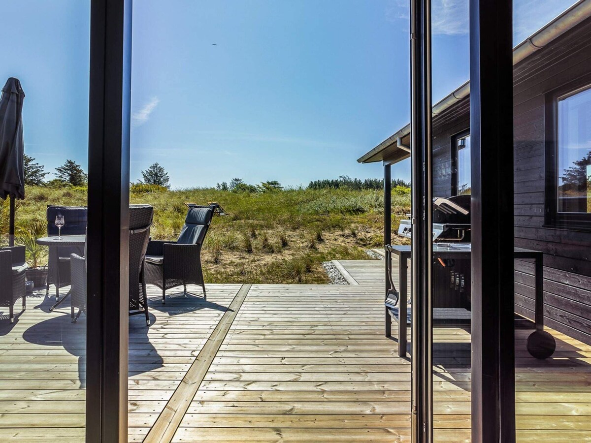 A wooden terrace is visible through large glass doors, featuring outdoor seating and a grill. Natural dunes are seen in the background under a clear blue sky, offering a peaceful connection to the surrounding landscape.