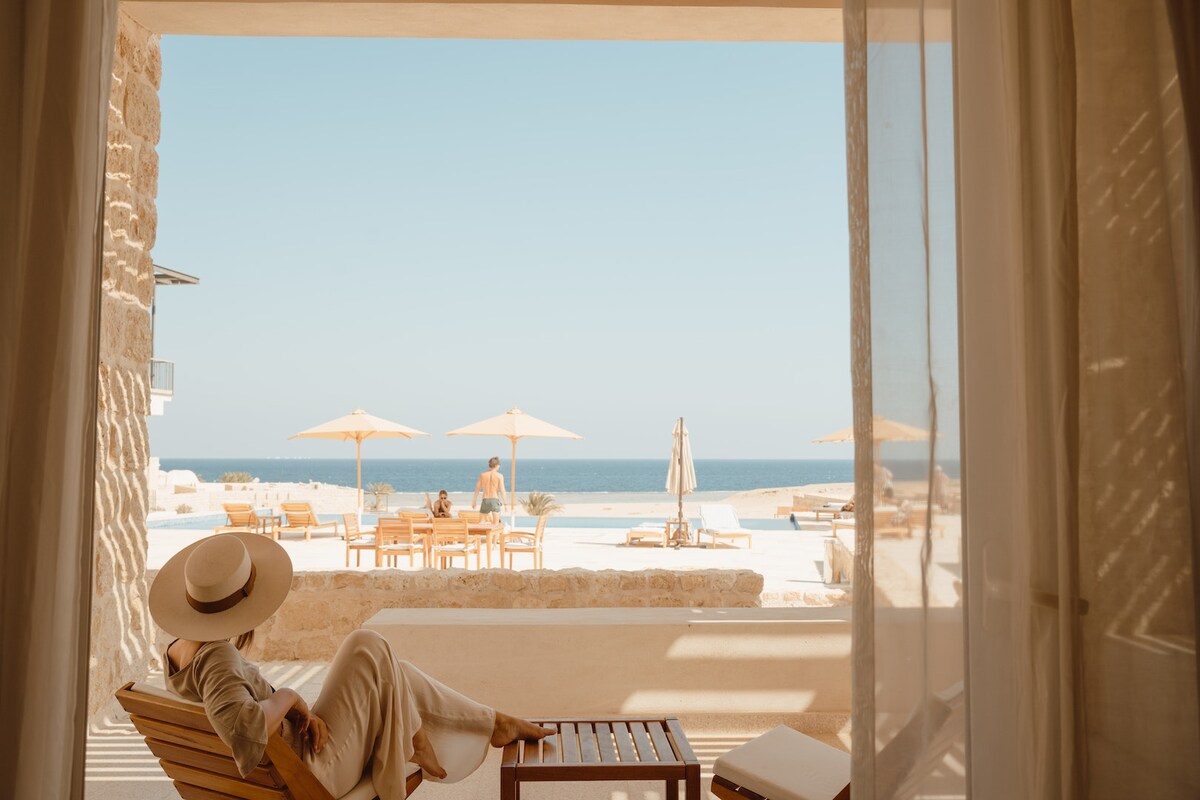 A spacious terrace offers a clear view of the outdoor swimming pool and the ocean beyond. Sun loungers are arranged around the pool, shaded by large umbrellas. A person relaxes in a wooden chair, wearing a wide-brimmed hat, while light filters through soft sheer curtains.