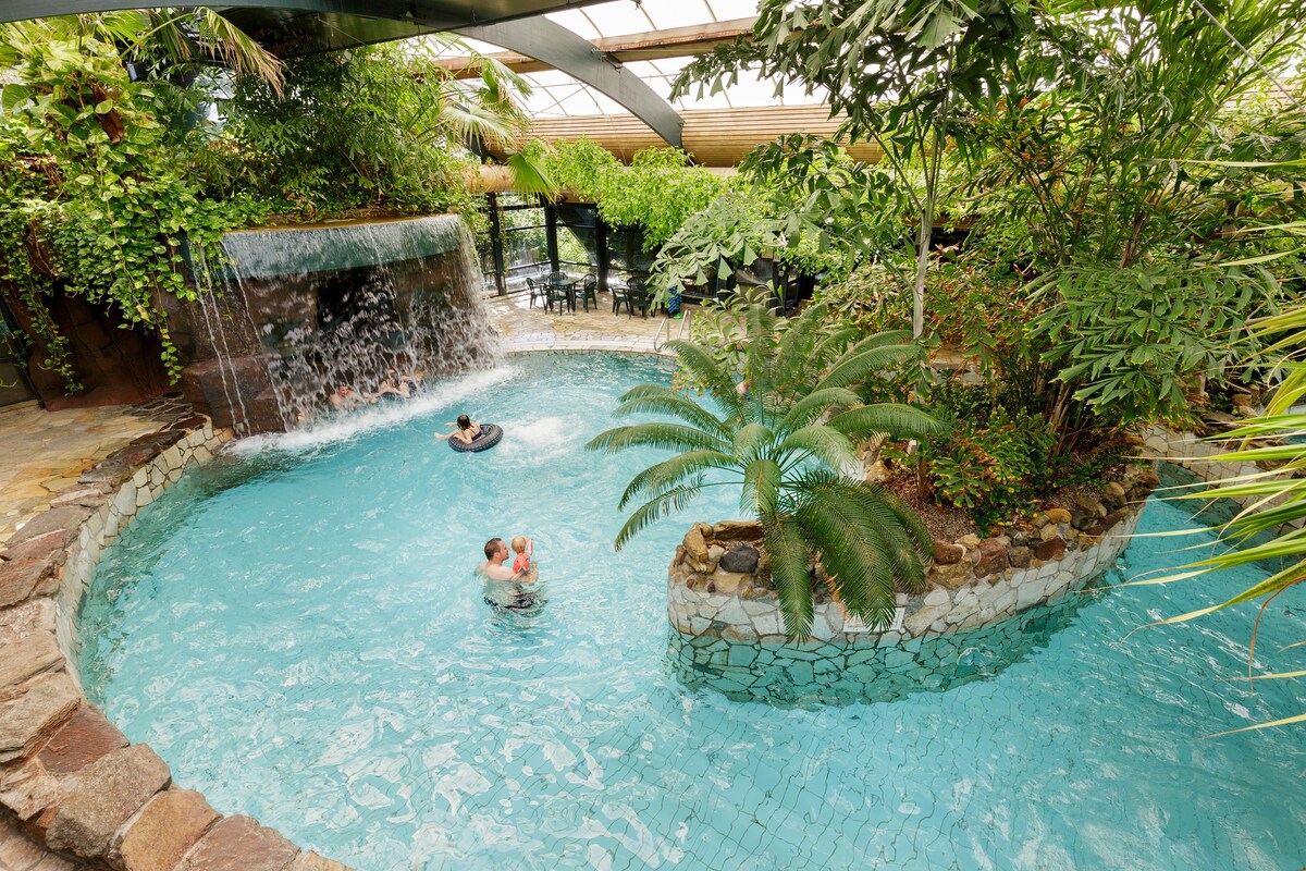 An indoor tropical pool features a cascading waterfall surrounded by lush greenery. Guests can be seen enjoying the water in an inviting atmosphere. The curved pool is bordered by natural stone and plants, enhancing the serene environment.