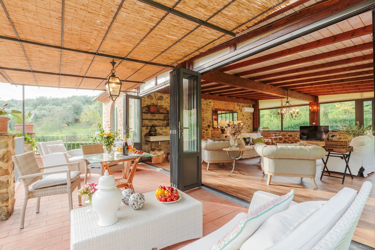 An open terrace area features a blend of comfortable seating arrangements with natural wicker furniture, a coffee table adorned with fresh fruit, and wooden beams. Expansive glass doors provide access to lush greenery beyond, while an inviting ambiance is created by decorative light fixtures.