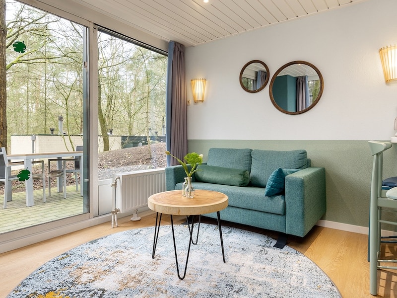 A cozy living area features a green sofa paired with a round wooden coffee table atop a patterned rug. Wall-mounted mirrors and warm light fixtures complement the space. Large windows allow for natural light and a view of the surrounding nature.