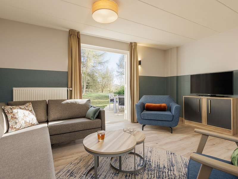 A renewed living room is presented with a sitting area including a gray sofa, a blue chair, and a round wooden table. A flatscreen TV is positioned on a media unit, while large glass doors lead to a terrace with outdoor seating.