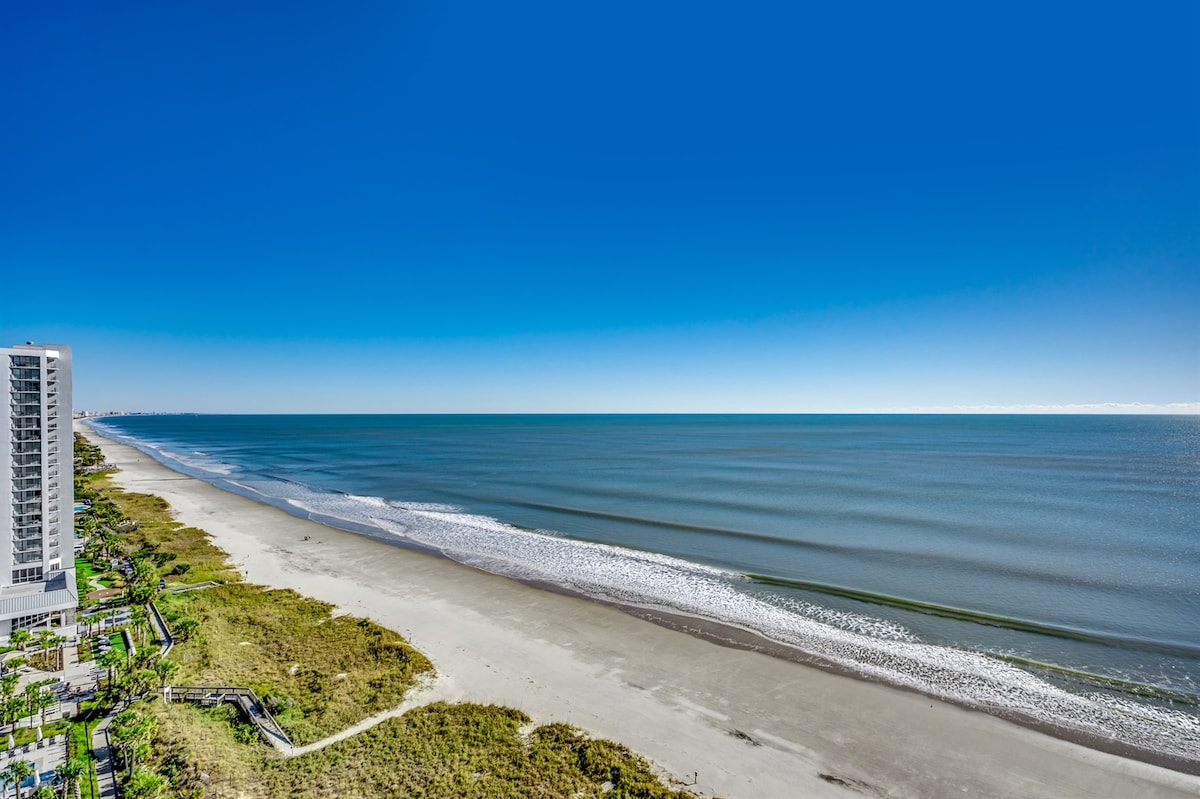 A serene ocean view captures the expansive shoreline with soft, rolling waves. The gentle sweep of the beach is visible, bordered by lush greenery. Clear blue skies stretch overhead, enhancing the tranquil scene of sandy shores and calm waters.