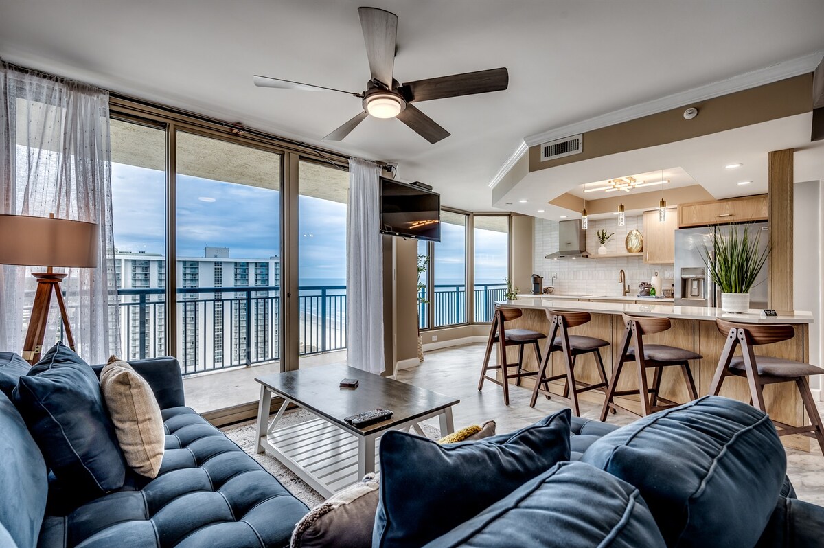 An open-concept living area features a comfortable sectional sofa facing a large window with ocean views. A dining area with wooden chairs complements a fully equipped kitchen, illuminated by natural light. Ceiling fans enhance air circulation within this modern space.