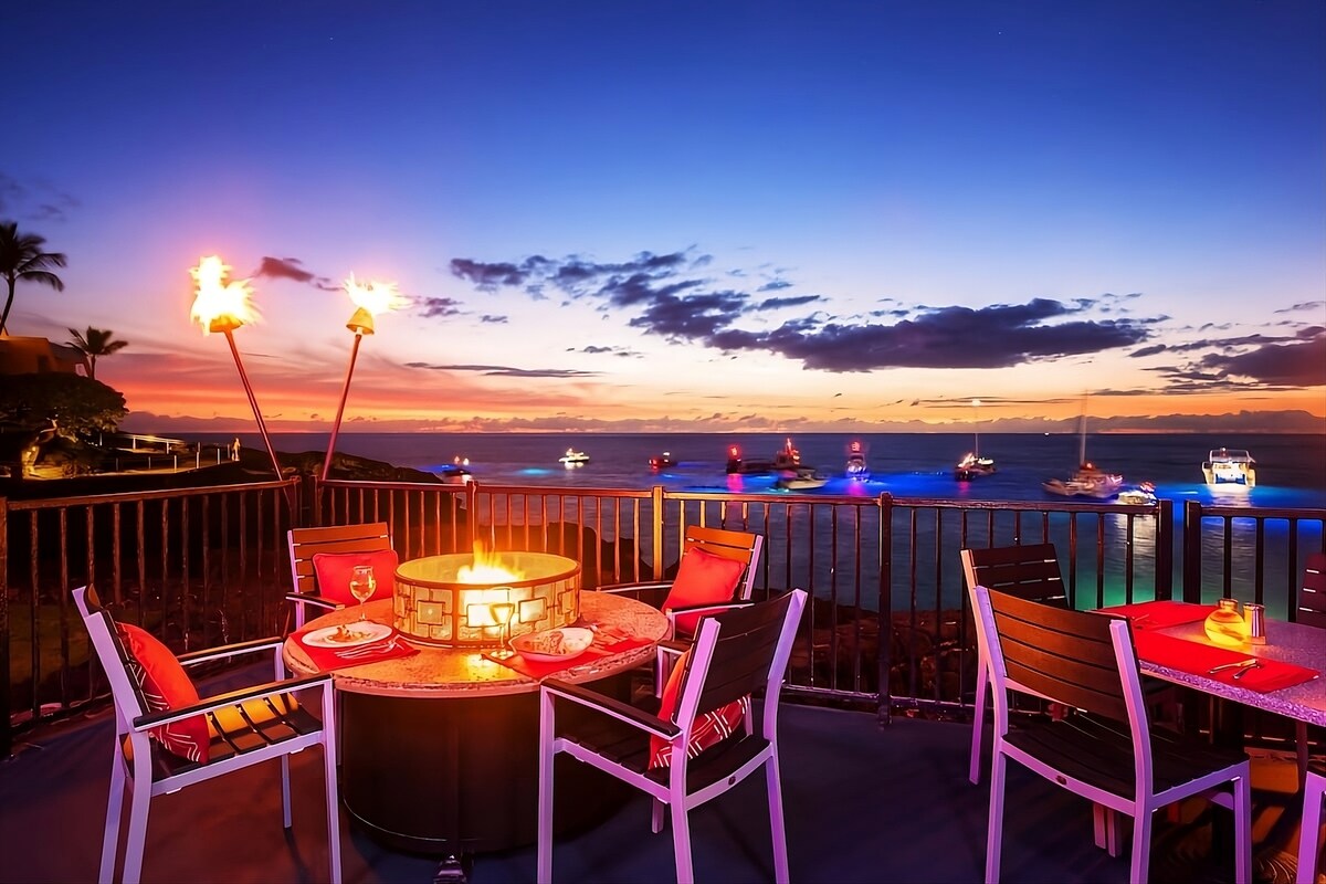 An outdoor dining area showcases a fire pit surrounded by dining tables with comfortable chairs. The vibrant sunset illuminates the ocean, enhancing the scenic atmosphere, with boats visible in the distance. Tiki torches flicker softly against the twilight sky.