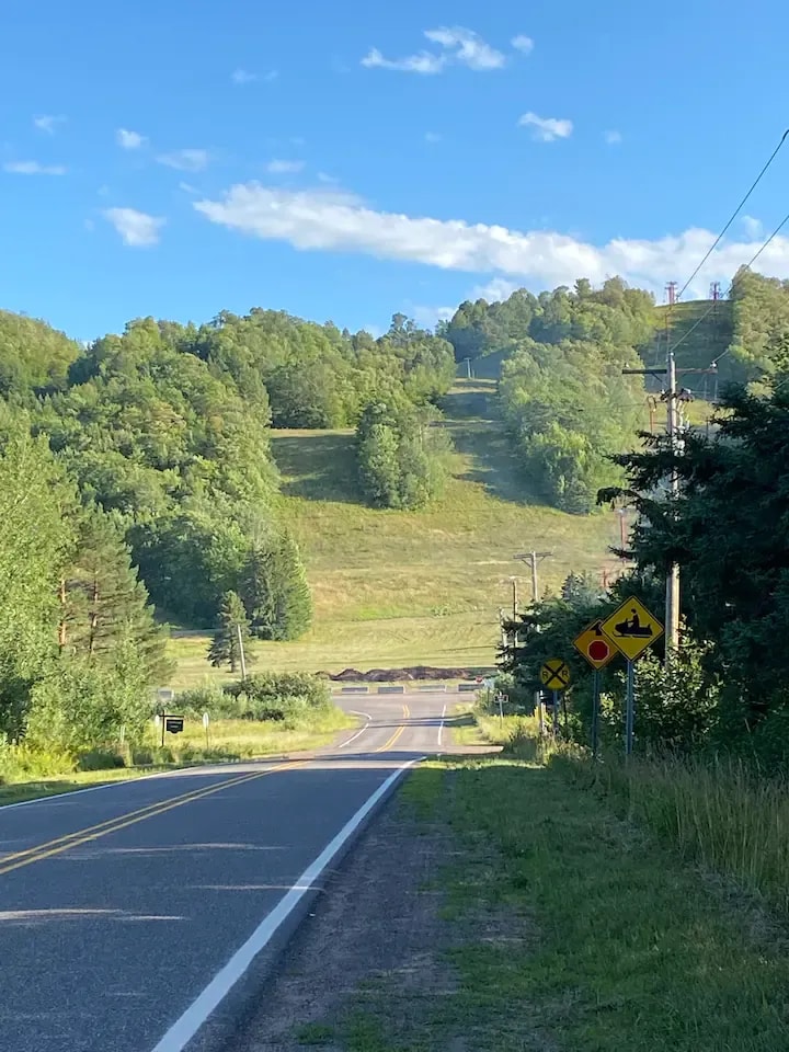 A winding road stretches through a green landscape, flanked by tall trees on either side. A ski slope is visible in the distance, nestled among rolling hills under a clear blue sky. Road signs alert drivers to curves ahead.