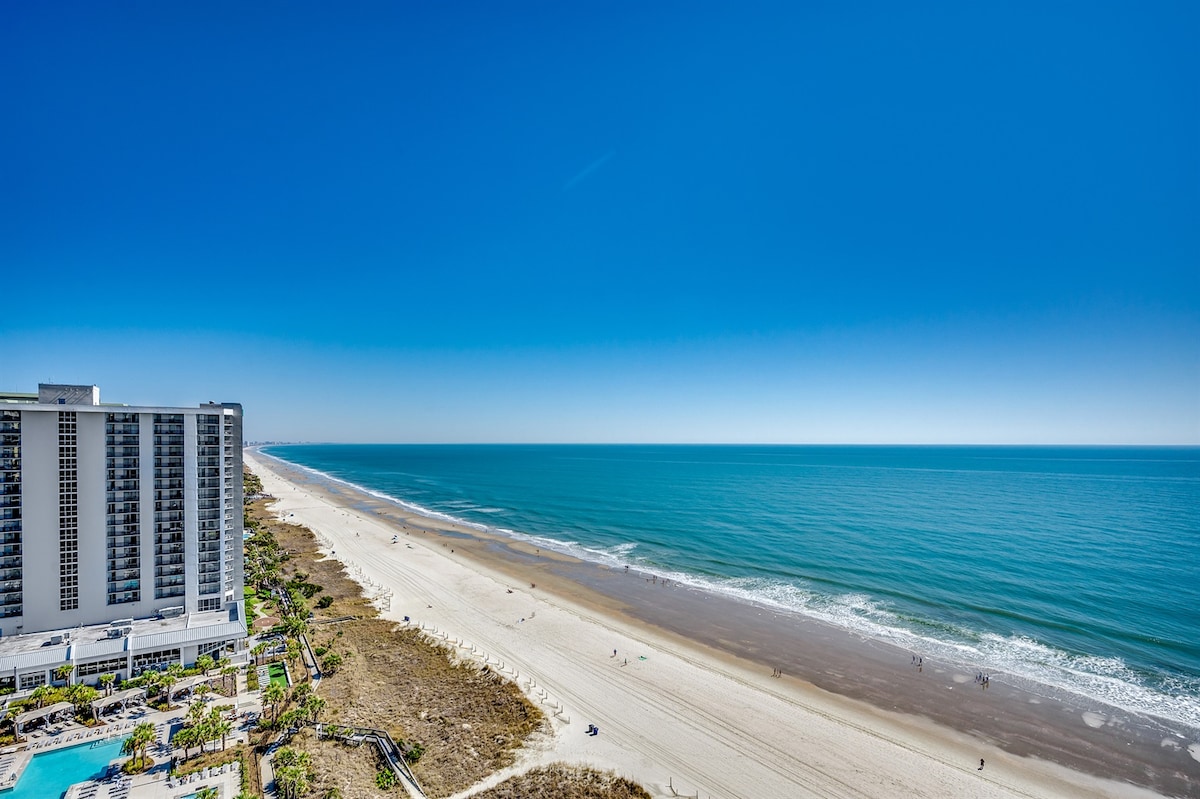An expansive view of the ocean is showcased, with soft sandy beaches stretching along the shoreline. The water glistens under a clear blue sky, while small figures can be seen enjoying the beach. A nearby building is visible, complementing the serene coastal scene.