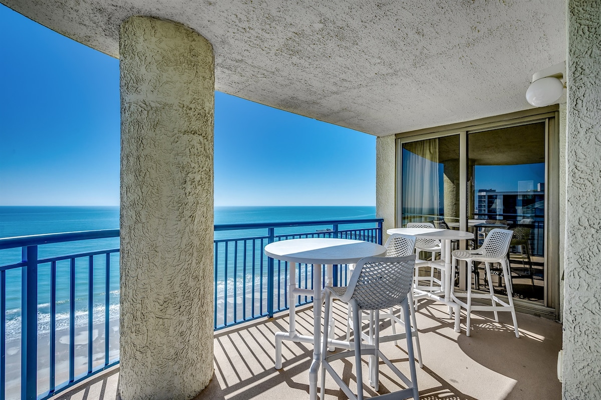A balcony overlooks the ocean, featuring white outdoor furniture including a round table and high-backed chairs. The expansive view captures the serene blue waters and clear sky, while light streaming through large glass doors brightens the space.