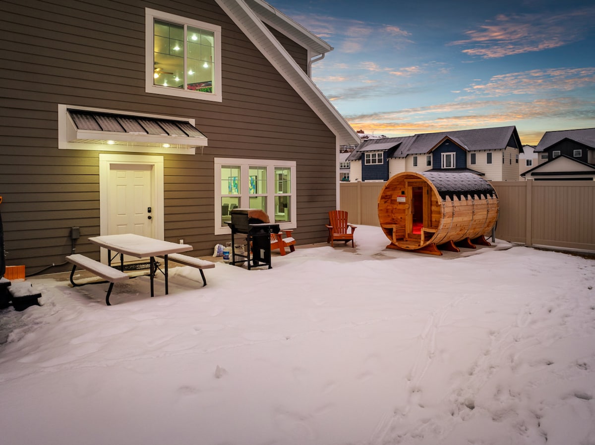 A snowy backyard features a picnic table and a gas grill, alongside a wooden barrel sauna. The home’s exterior is comprised of grey siding with large windows, allowing light to spill into the space. The evening sky displays soft colors at sunset.