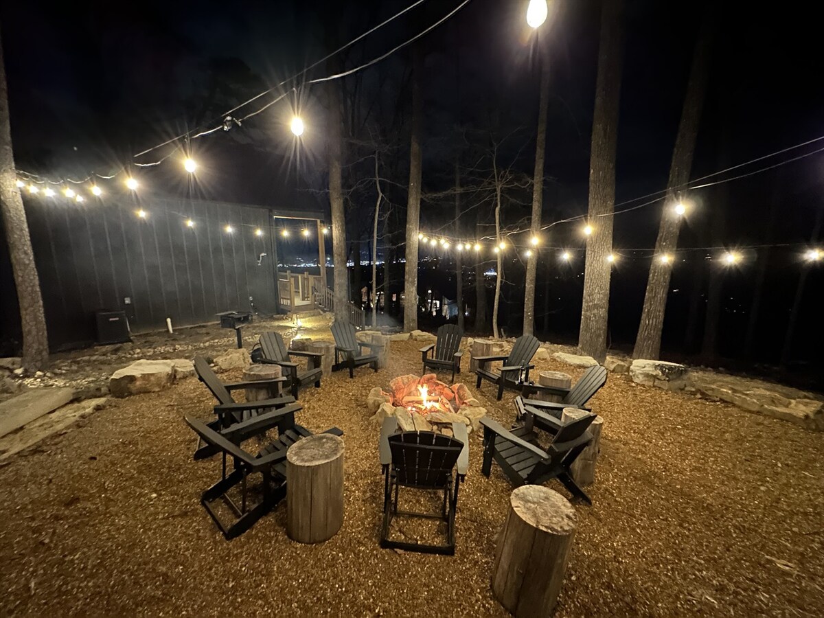 An outdoor fire pit area is surrounded by wooden adirondack chairs on a gravel surface. Soft lighting from hanging bulbs creates a warm ambiance among the trees. The night sky is visible, and a view of distant lights adds to the inviting setting.