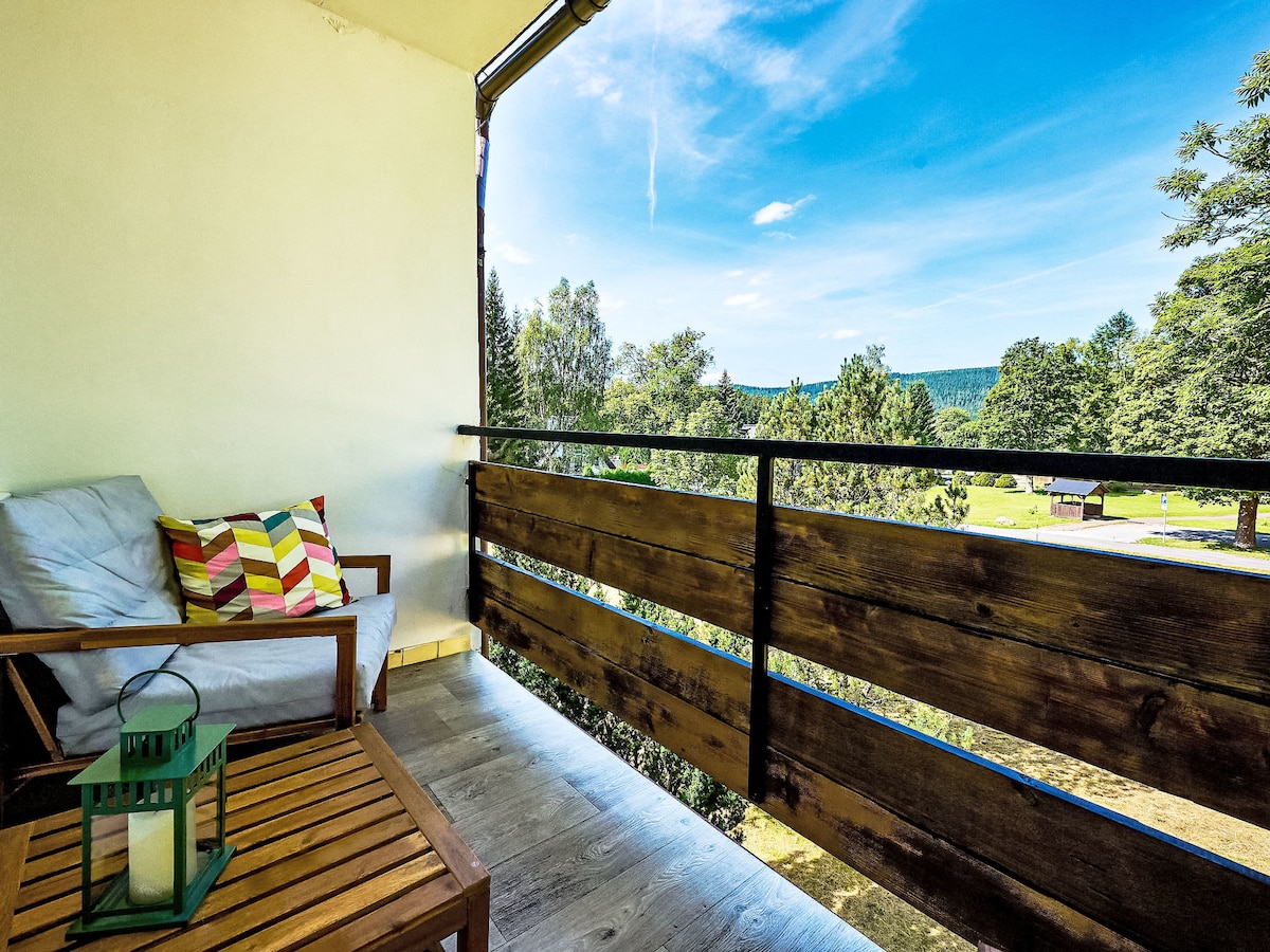 A balcony is featured with a wooden railing and views of the surrounding landscape. A comfortable armchair with decorative cushions is positioned beside a wooden table, complemented by a lantern. Bright blue skies and greenery enhance the outdoor space.