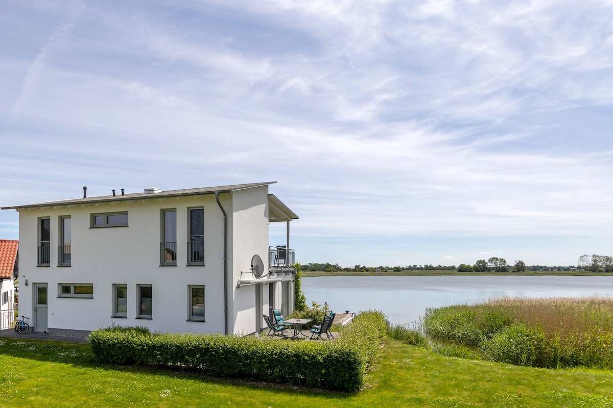 The modern two-story building is set along the water's edge, facing a calm body of water. The exterior features a clean design with large windows and a balcony. Lush green grass surrounds the property, enhancing the natural setting.