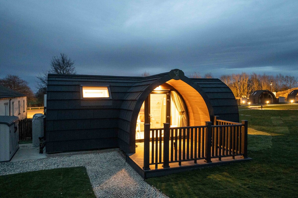 A modern glamping pod is illuminated at dusk, displaying a welcoming entrance with a wooden deck and fence. The curved roof and sleek exterior harmonize with the serene surroundings, while soft light glows from the interior, highlighting the cozy and inviting atmosphere.