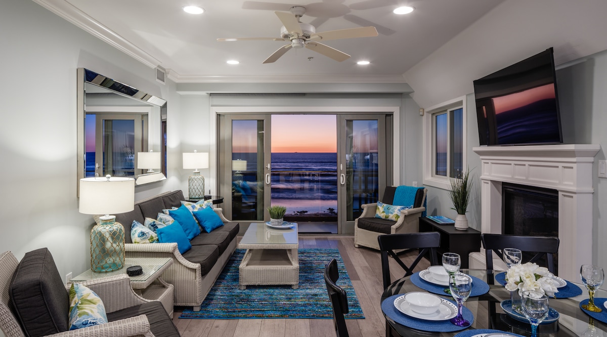 A spacious living area is furnished with a mix of light-colored sofas and a coffee table, creating a relaxed setting. Large sliding glass doors open to reveal expansive ocean views, while evening light softly illuminates the room. A dining table is set with dishes, accommodating a welcoming atmosphere.