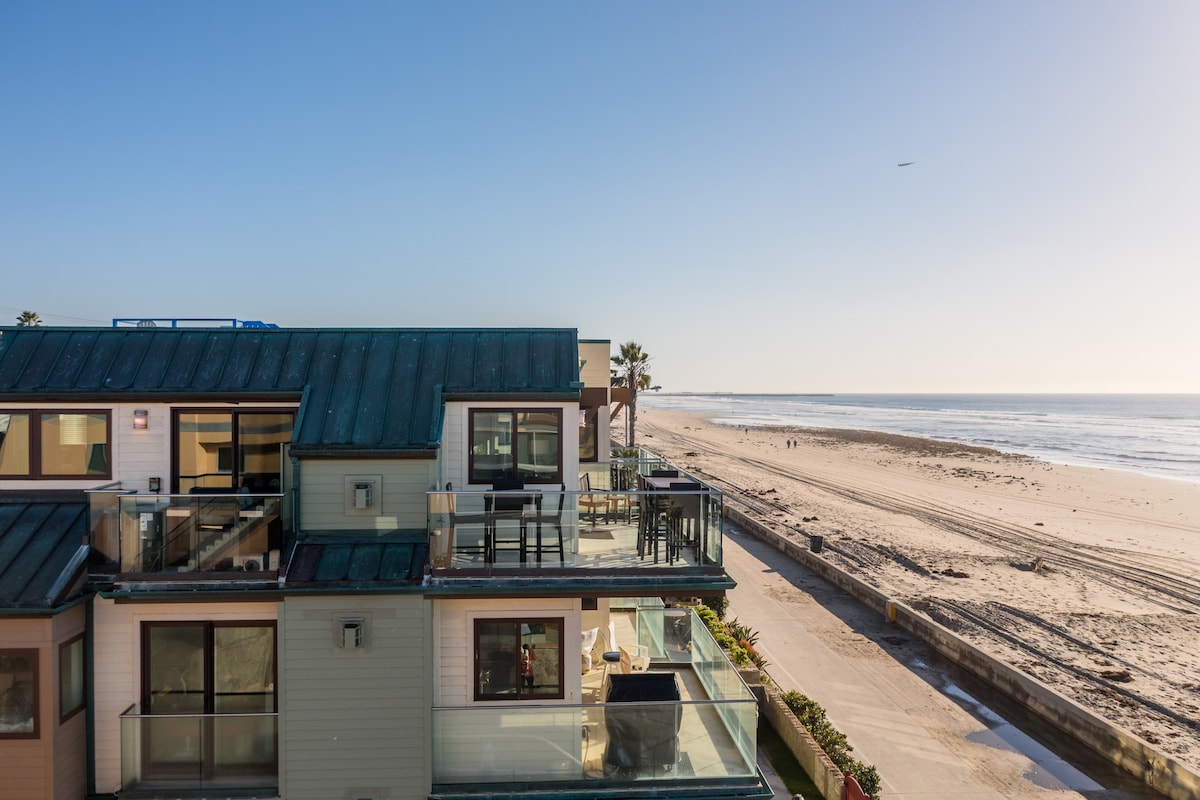An aerial view showcases the penthouse condo, featuring multiple outdoor balconies with clear views of the sandy beach and ocean. The coastline stretches along the horizon, bordered by palm trees, while the boardwalk invites leisurely strolls beside the water.
