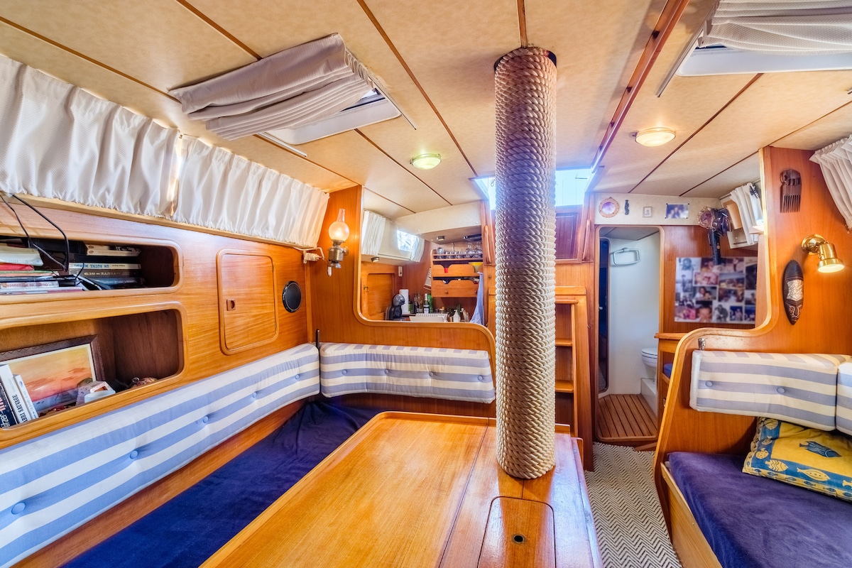 The interior space features warm wooden paneling with cozy seating along the walls. A dining table is positioned centrally, surrounded by upholstered cushions. Warm lighting and a ventilation system are present, contributing to a comfortable atmosphere. A doorway leads to a well-appointed bathroom.