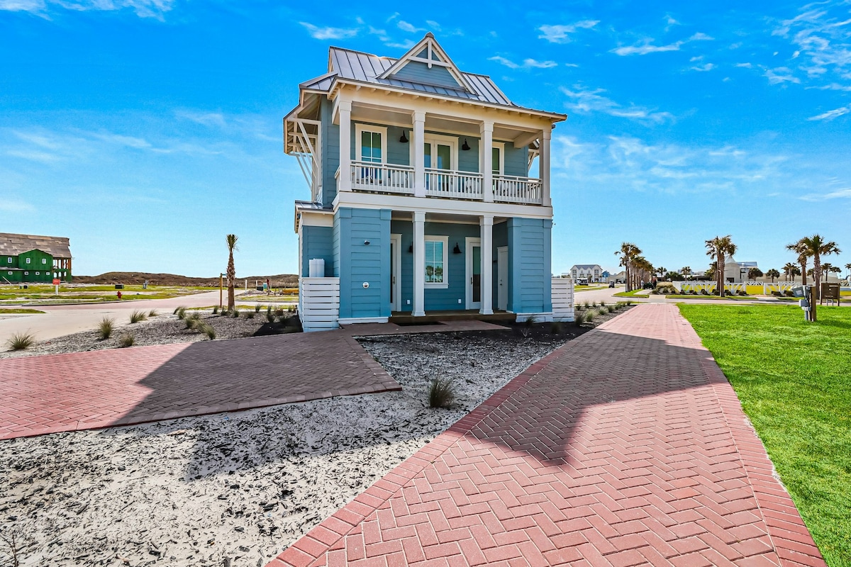 A two-story coastal home features a light blue exterior with white trim and balconies on both levels. Paved walkways lead to the entrance, framed by landscaped areas with sparse vegetation. Palm trees are visible nearby, suggesting a tropical environment.
