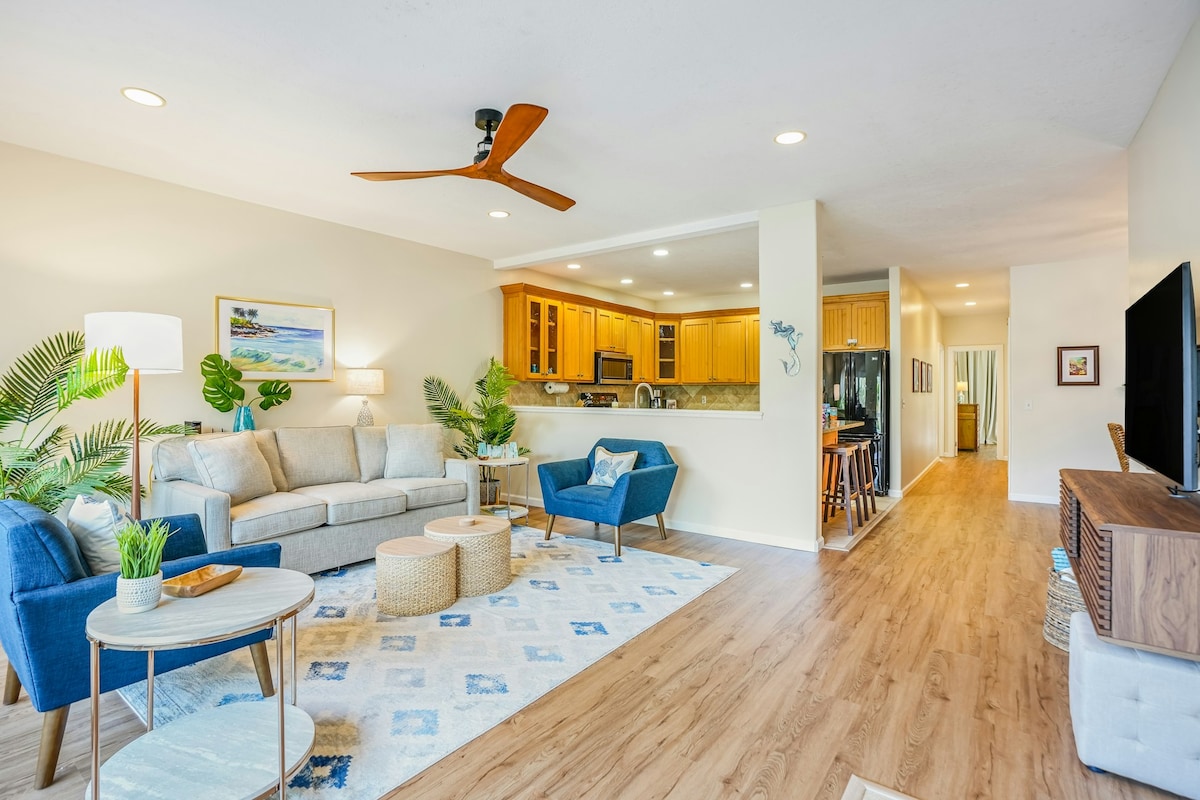 The living area features a spacious layout with a light grey sofa and two vibrant blue chairs. A circular coffee table rests on a textured area rug. The kitchen is partially visible, showcasing wooden cabinetry and modern appliances. Natural light fills the room, enhancing a welcoming atmosphere.