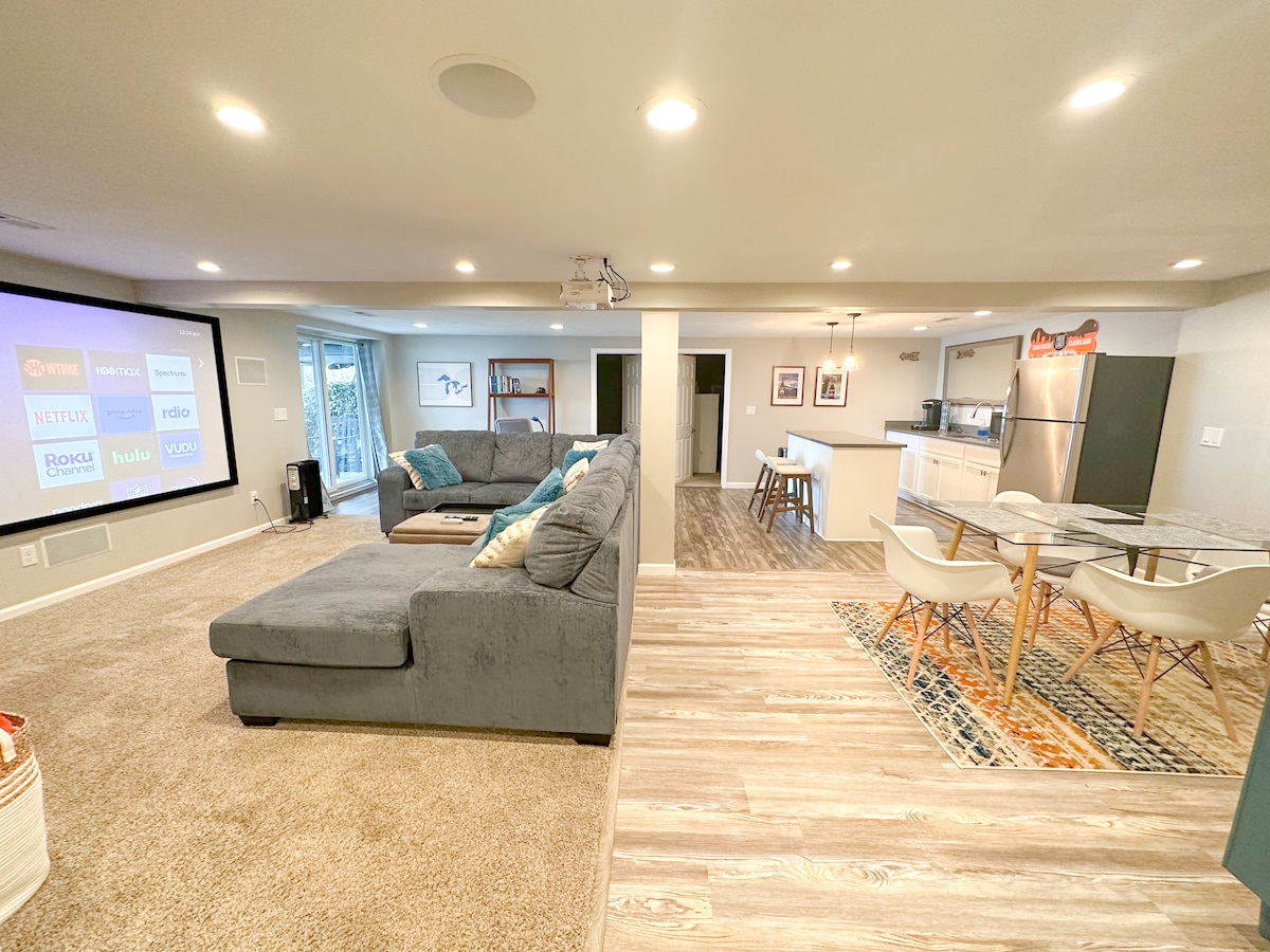 The spacious lower level features a comfortable seating area with a large sectional couch, paired with a contemporary dining table. A projection screen dominates one wall, while a well-equipped kitchen area is visible, showcasing modern appliances and an open layout, enhancing usability for gatherings.